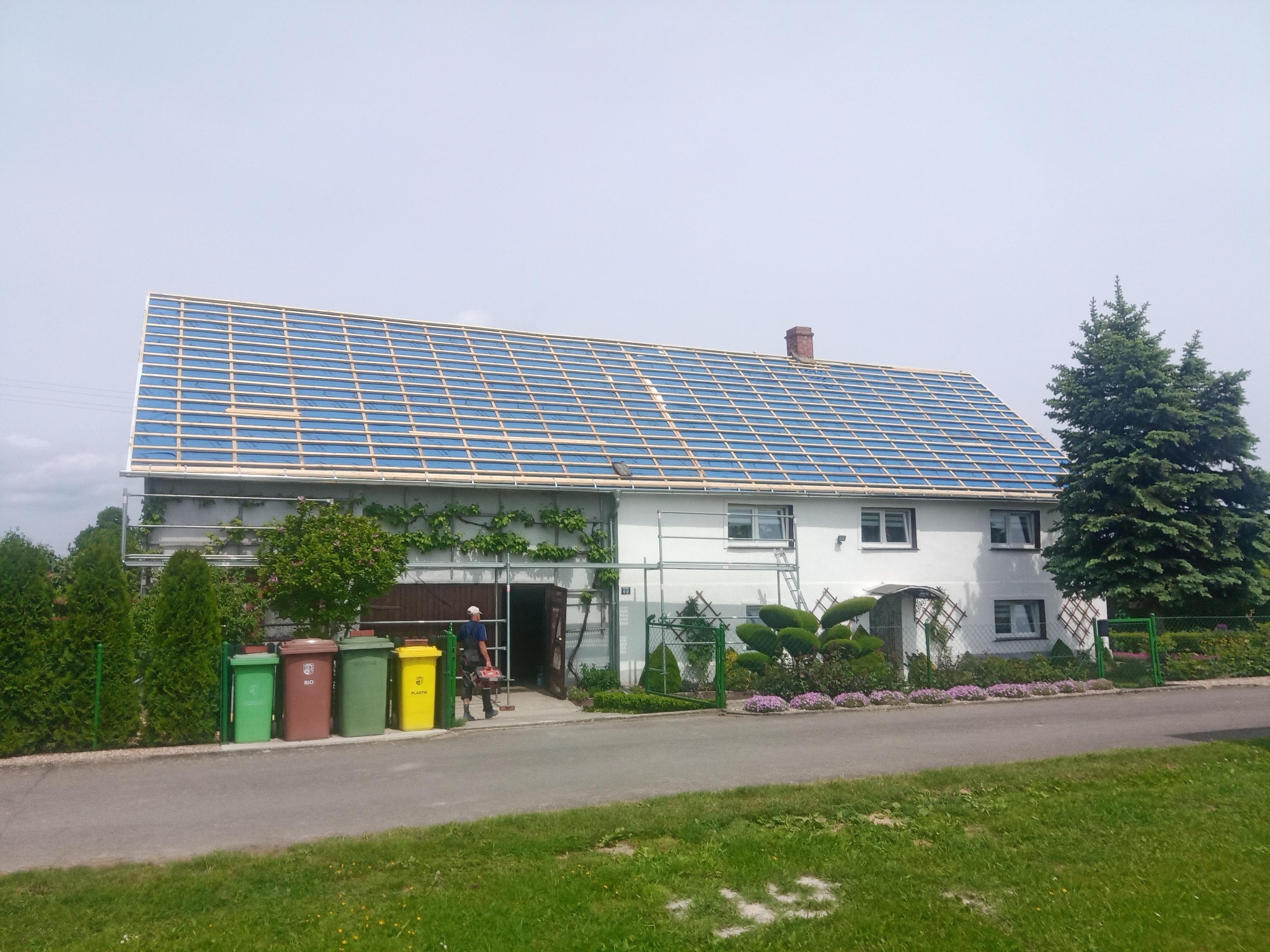 Dom jednorodzinny z odsłoniętą konstrukcją dachu pokrytą niebieską folią, widoczny pracownik wchodzący do budynku z narzędziami.