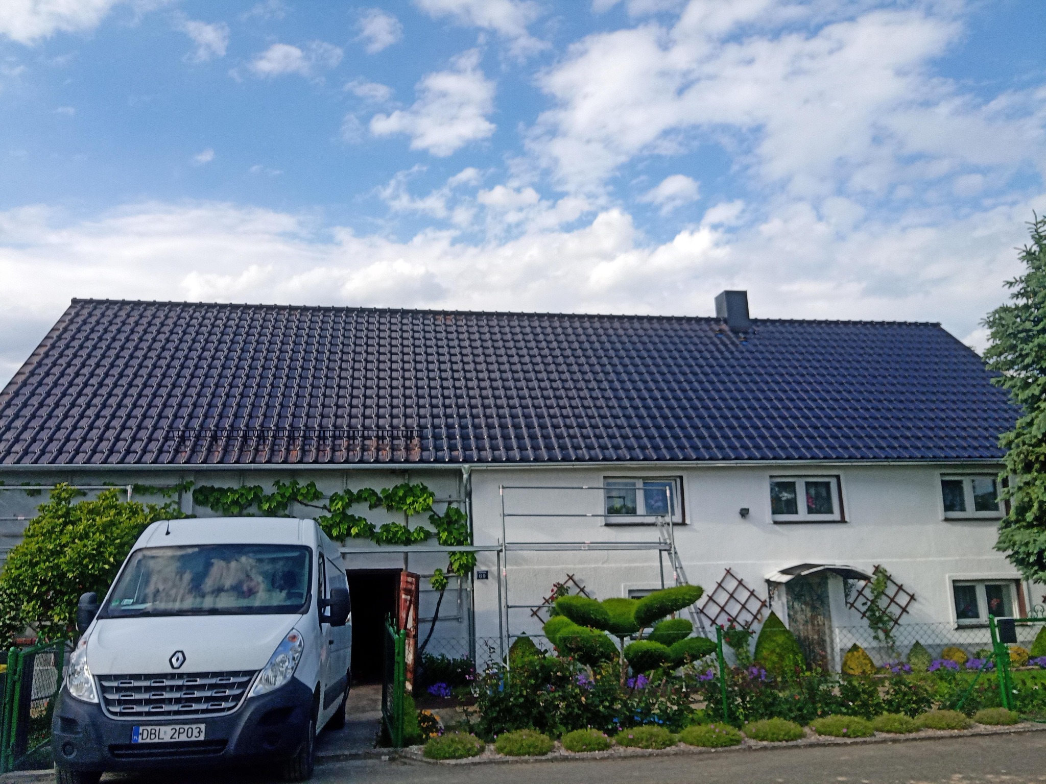 Dom z ciemną dachówką ceramiczną, widoczny biały bus Renault Master, rusztowanie przy ścianie i zadbany ogród z ozdobnymi krzewami.