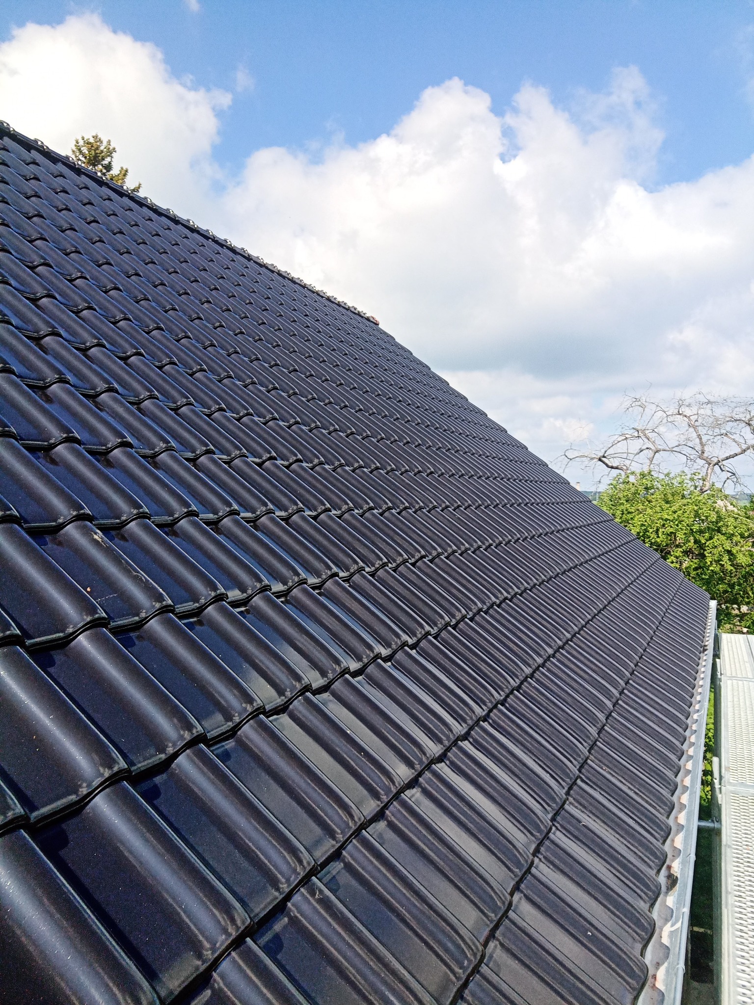 Ciemna dachówka ceramiczna ułożona na skośnym dachu, widok z dołu, z fragmentem rusztowania i błękitnym niebem w tle.