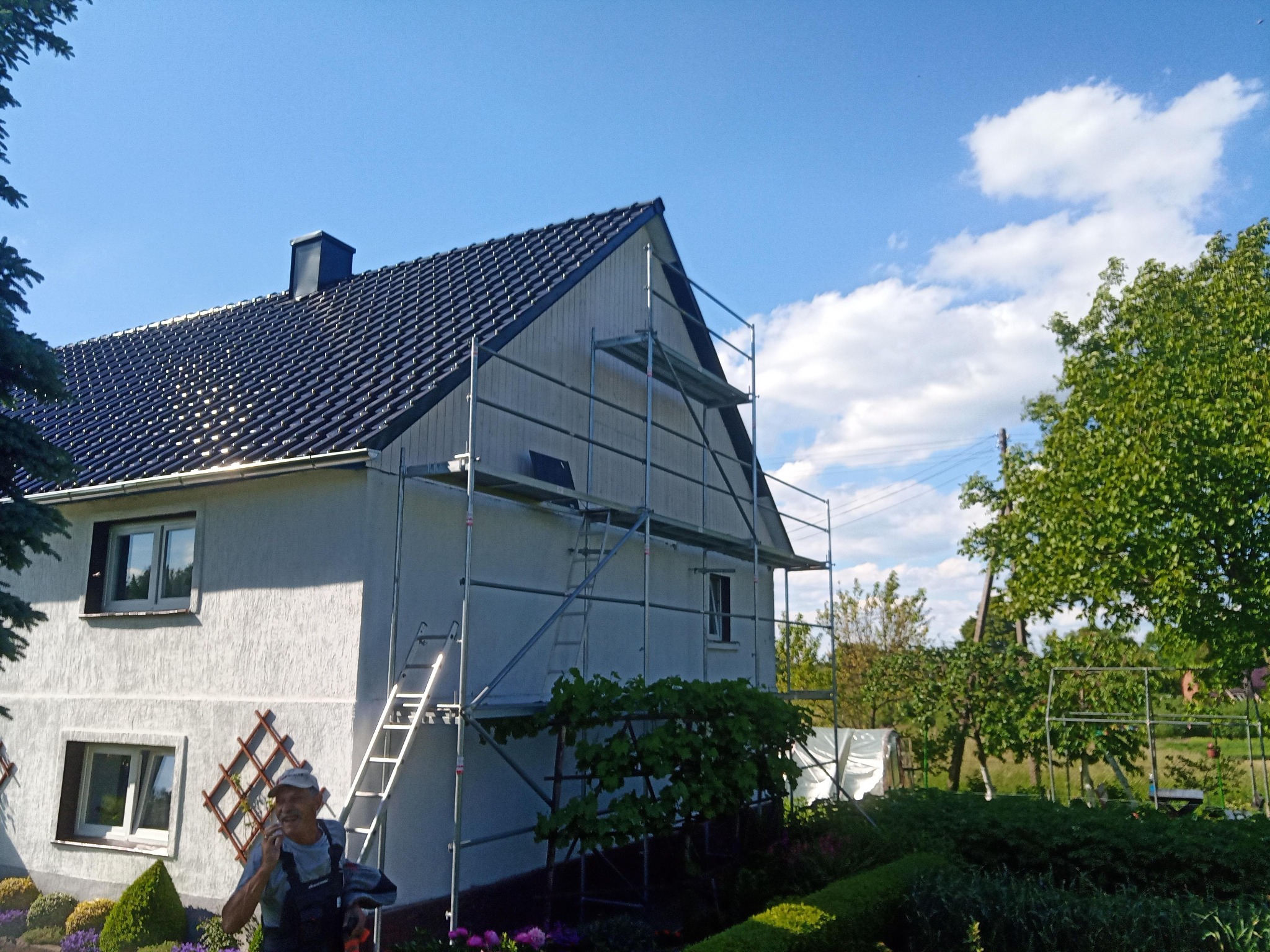 Dom jednorodzinny w trakcie prac z nowym, ciemnym dachem i rusztowaniem, mężczyzna w kombinezonie roboczym rozmawia przez telefon.