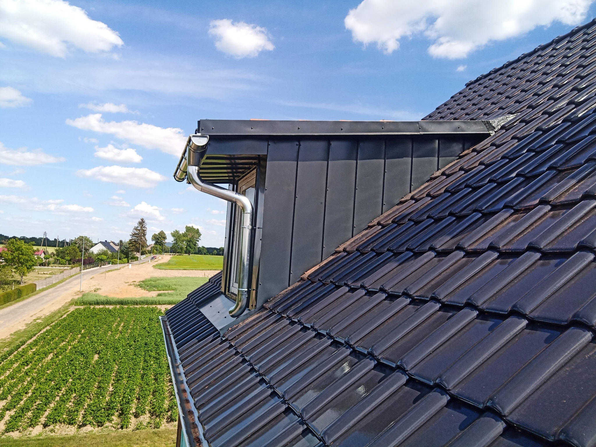 Widok z góry na fragment dachu z ciemnej dachówki ceramicznej, okno dachowe w obudowie z blachy i nową, błyszczącą rynnę spustową, na tle błękitnego nieba i krajobrazu wiejskiego.