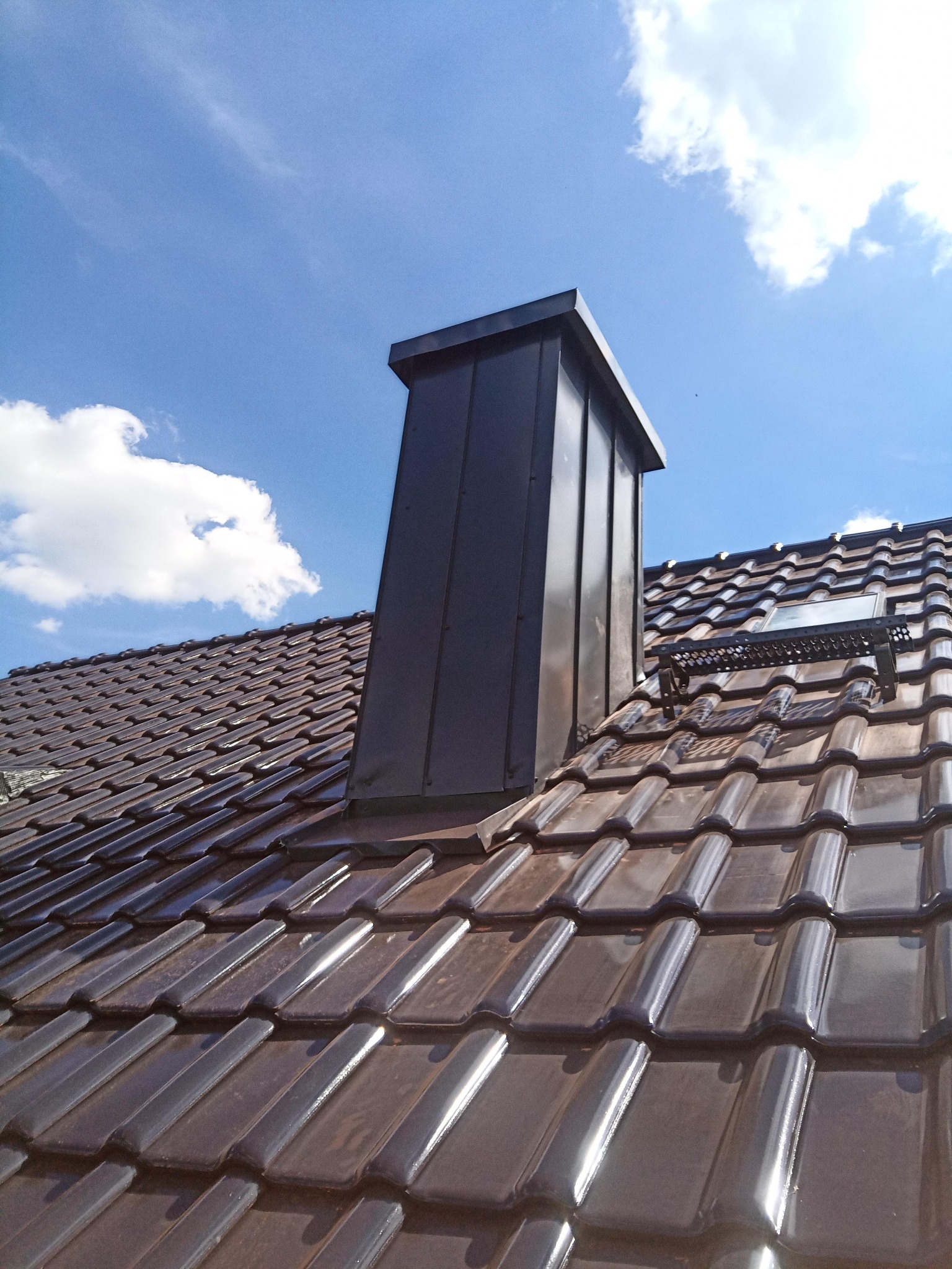 Nowy dach z brązowej dachówki ceramicznej, z oknem dachowym i metalowym kominem na tle błękitnego nieba z chmurami.
