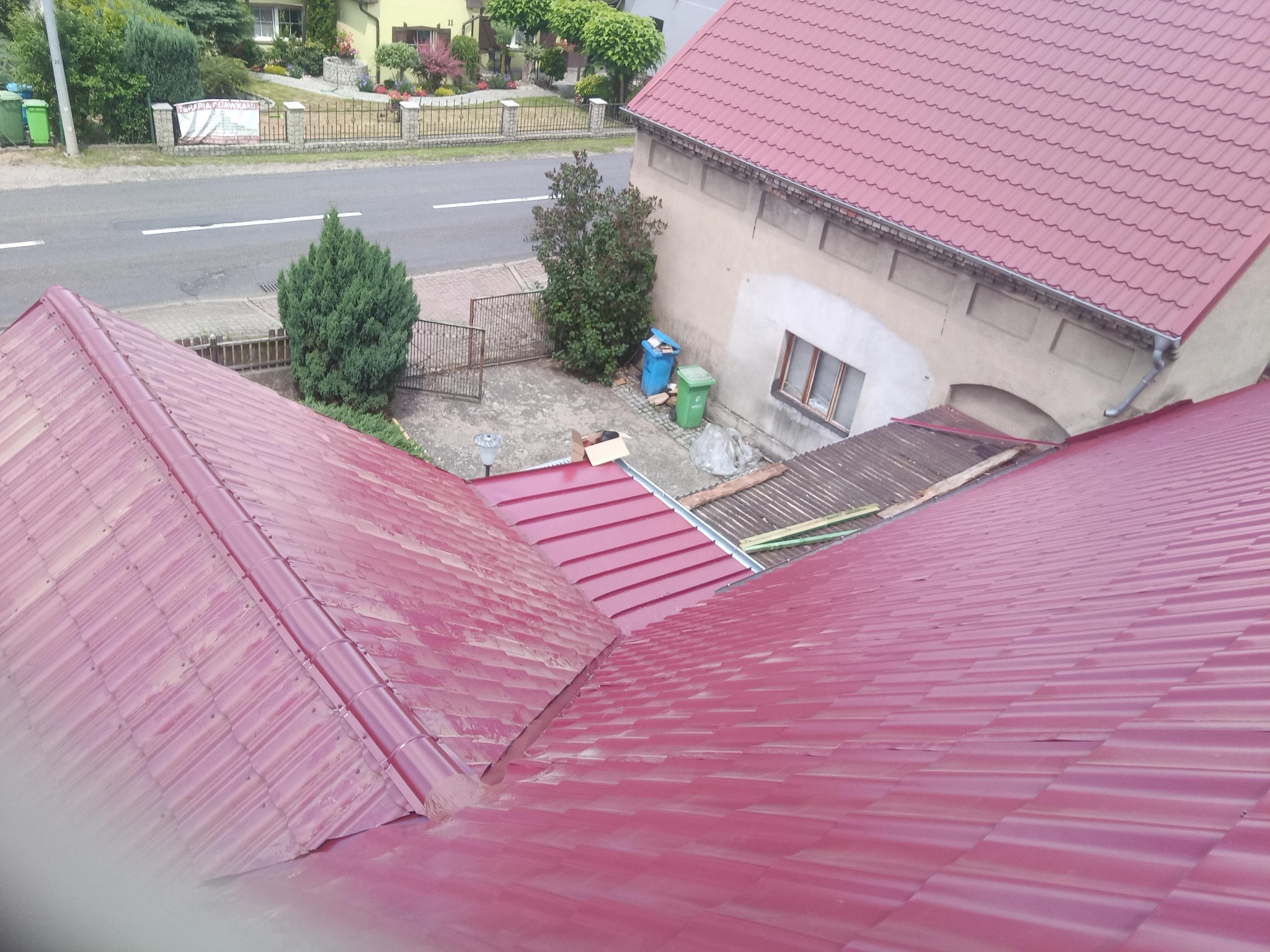 Widok z góry na fragment dachu z blachodachówki w kolorze bordowym, z widoczną kalenicą i fragmentem rynny, w tle budynek mieszkalny i otoczenie.
