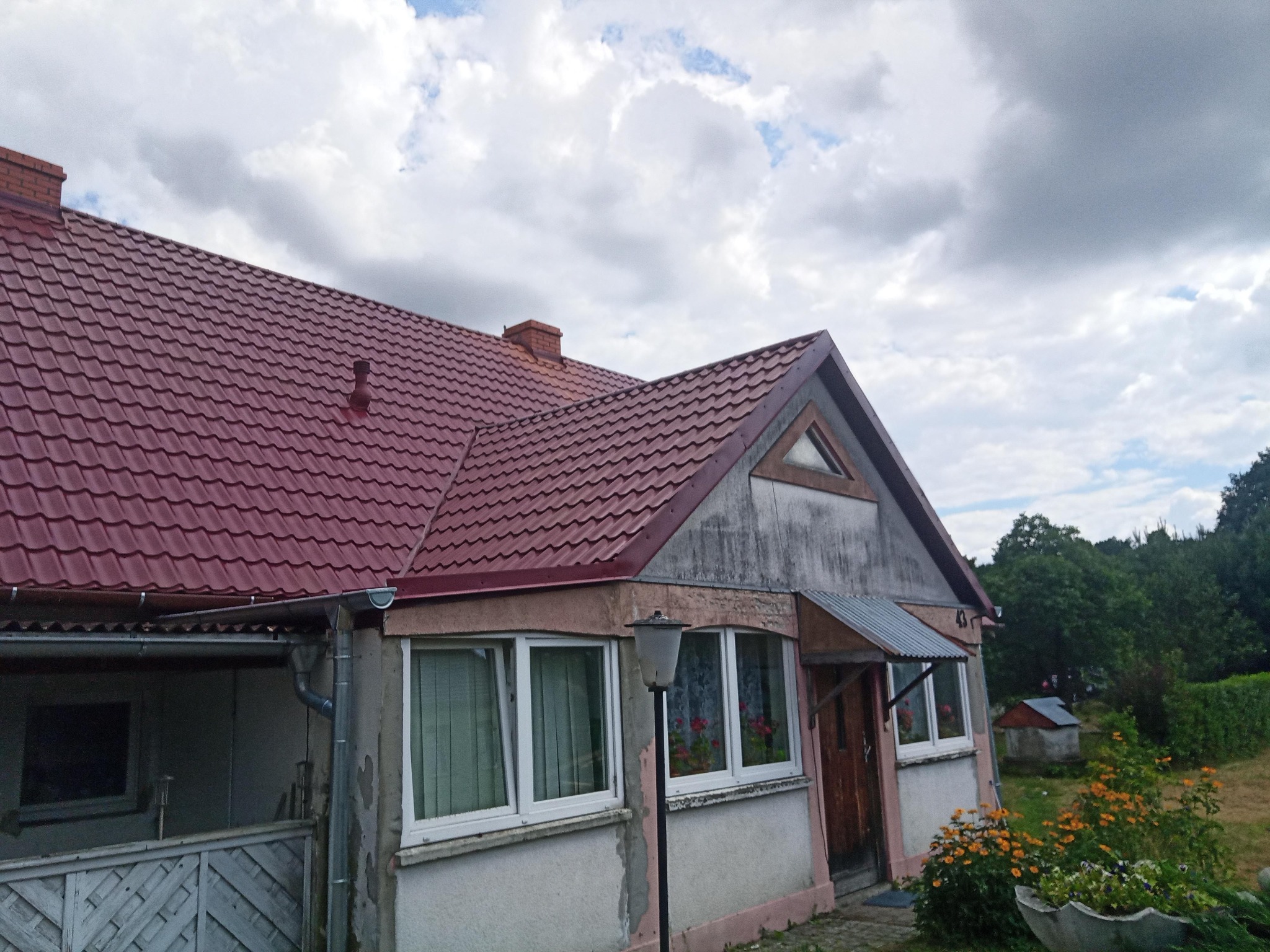 Dom jednorodzinny z nowym, bordowym dachem z blachodachówki, widoczne rynny i komin z cegły, na tle pochmurnego nieba.