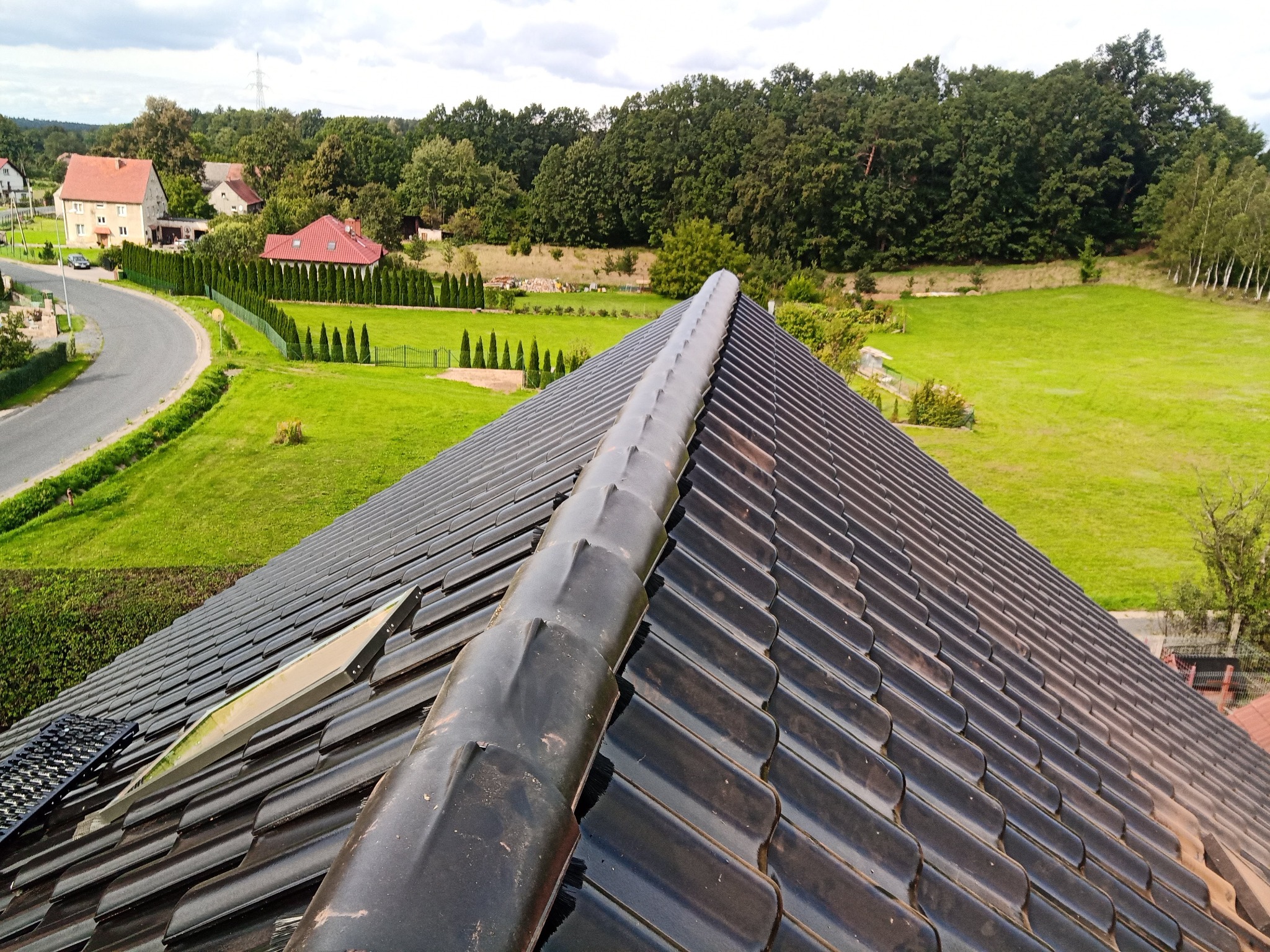 Widok z góry na ciemną dachówkę ceramiczną z widocznym systemem mocowania, w tle krajobraz wiejski z domami i polami.