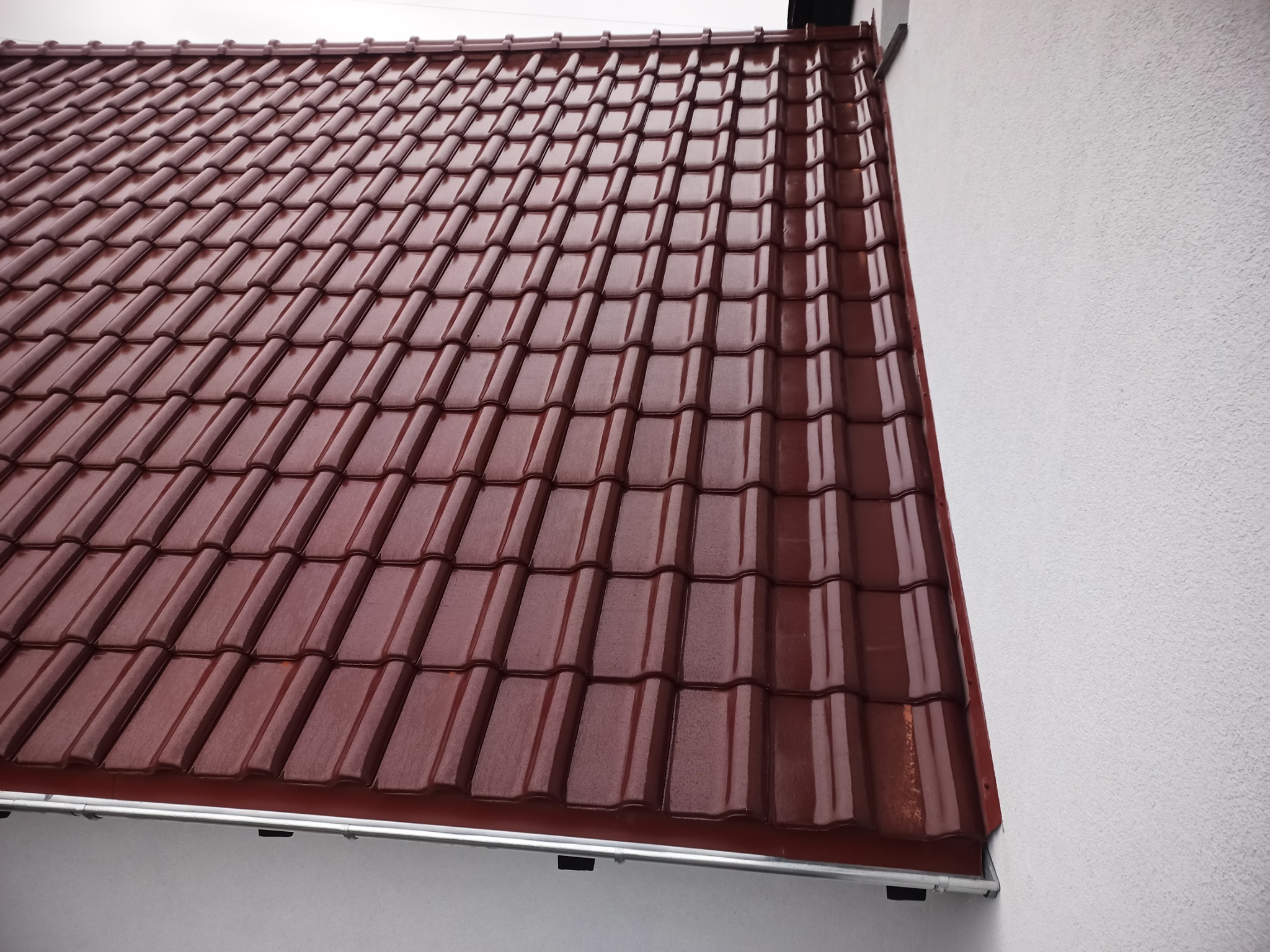 Czerwona dachówka ceramiczna na skośnym dachu, widoczna z bliska, z fragmentem białej ściany budynku po prawej stronie.
