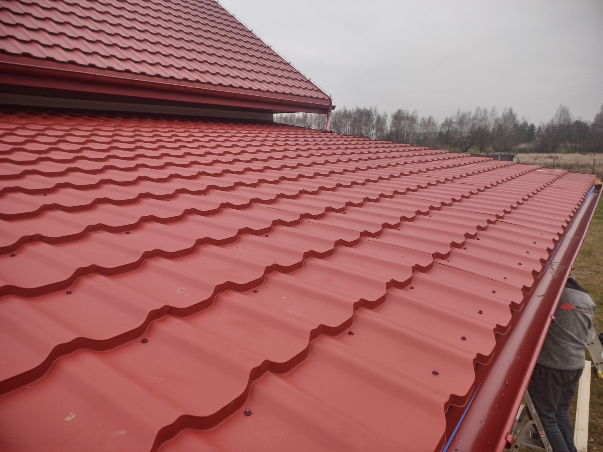 Czerwony dach z blachodachówki modułowej, widok z góry, z zamontowanymi rynnami w kolorze dachu i elementami mocującymi. W tle drzewa i pochmurne niebo.