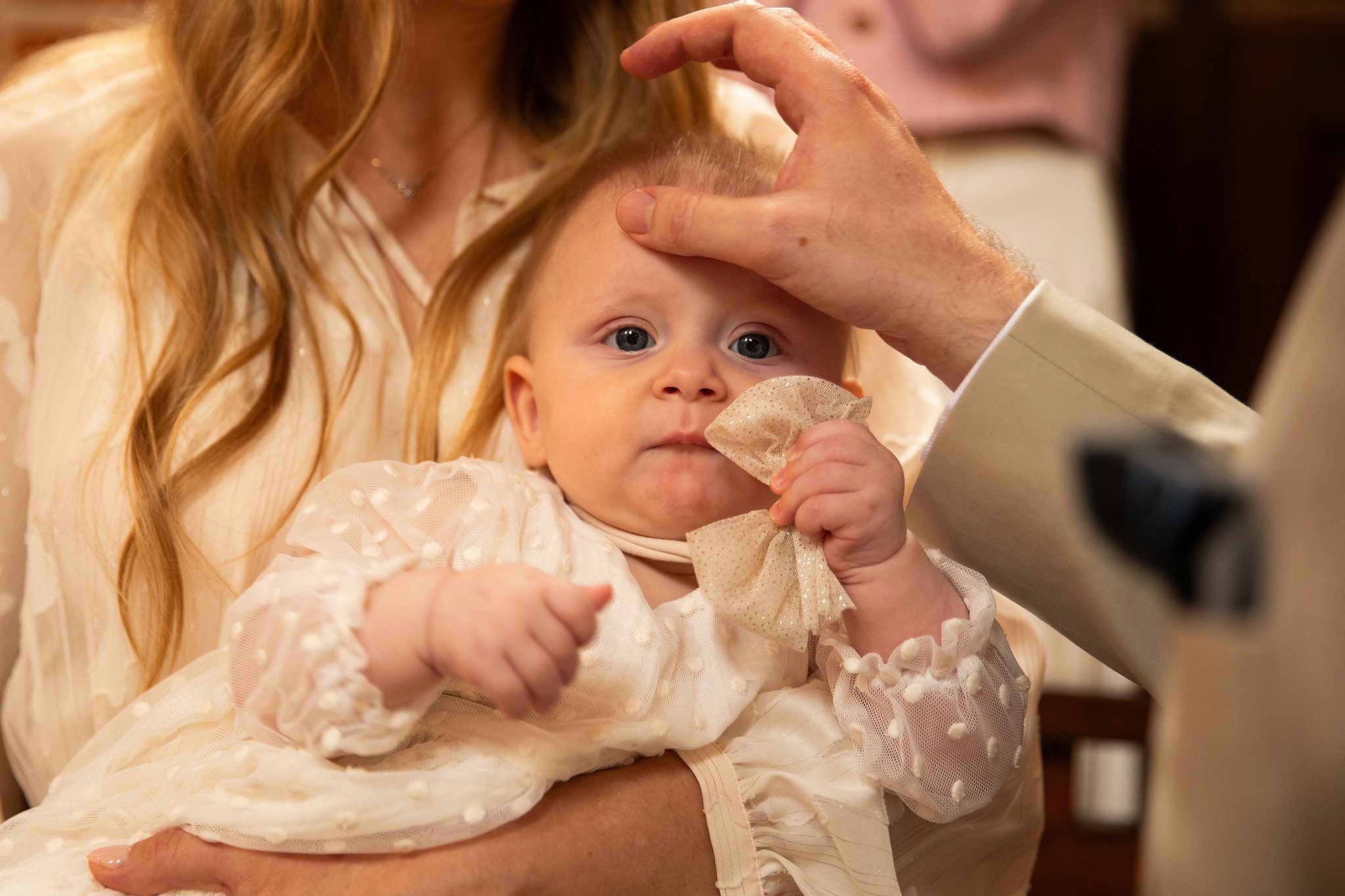 Chrzest dziecka: dłoń kapłana dotyka czoła niemowlęcia ubranego w białą szatkę z koronką, trzymanego przez kobietę. Ujęcie z bliska, ciepłe światło.