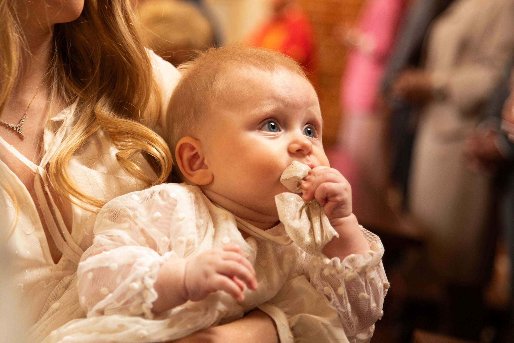 Urocze niemowlę w kremowej sukience z koronką, trzymane na rękach przez mamę. Dziecko z zaciekawieniem patrzy w górę, trzymając w rączce materiał. Delikatne, ciepłe światło.