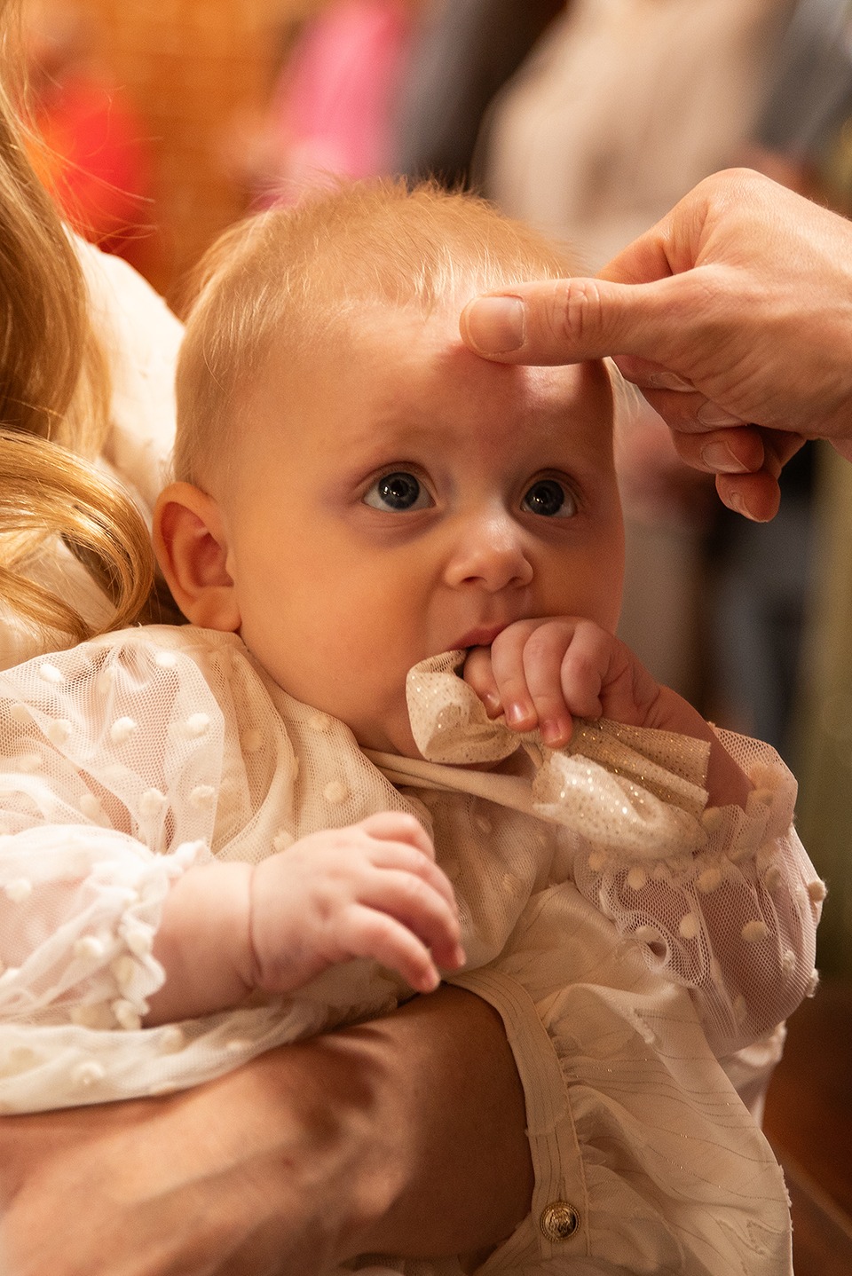 Chrzest: dłoń dotyka czoła niemowlęcia w białej, eleganckiej sukience. Dziecko trzyma w ustach ozdobną chusteczkę, spoglądając w górę. Ciepłe, naturalne światło.