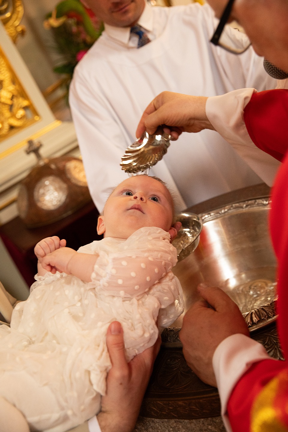 Chrzest niemowlęcia w kościele: ksiądz polewa wodą głowę dziecka trzymanego przez rodziców, w tle złote dekoracje i białe szaty.