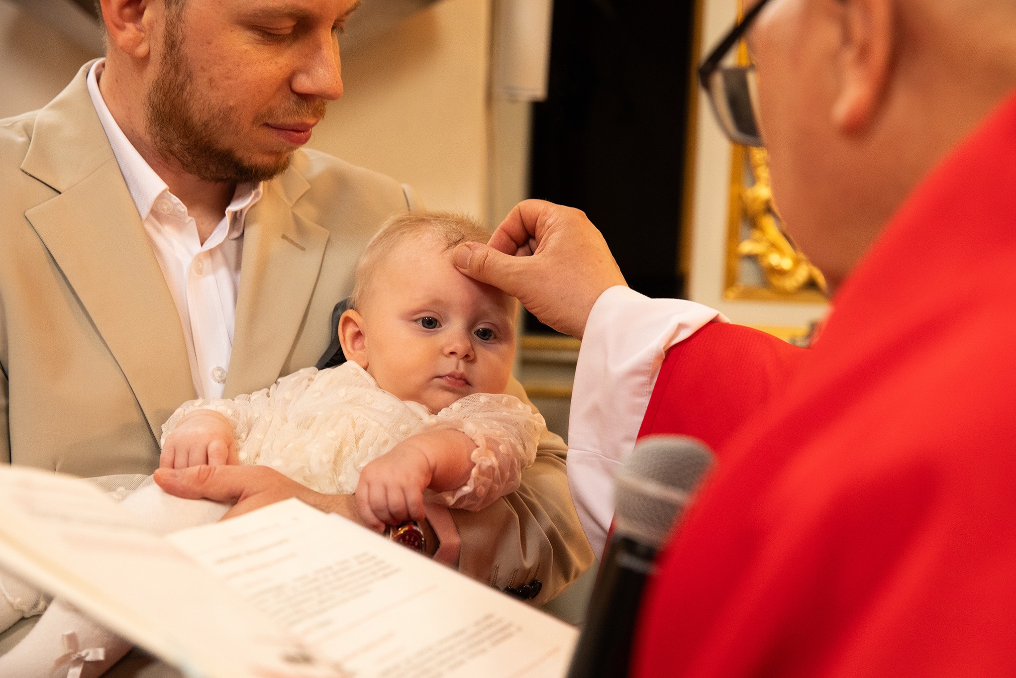 Chrzest dziecka: ksiądz w czerwonej szacie dokonuje namaszczenia czoła niemowlęcia trzymanego przez ojca w beżowym garniturze. W tle mikrofon i księga.