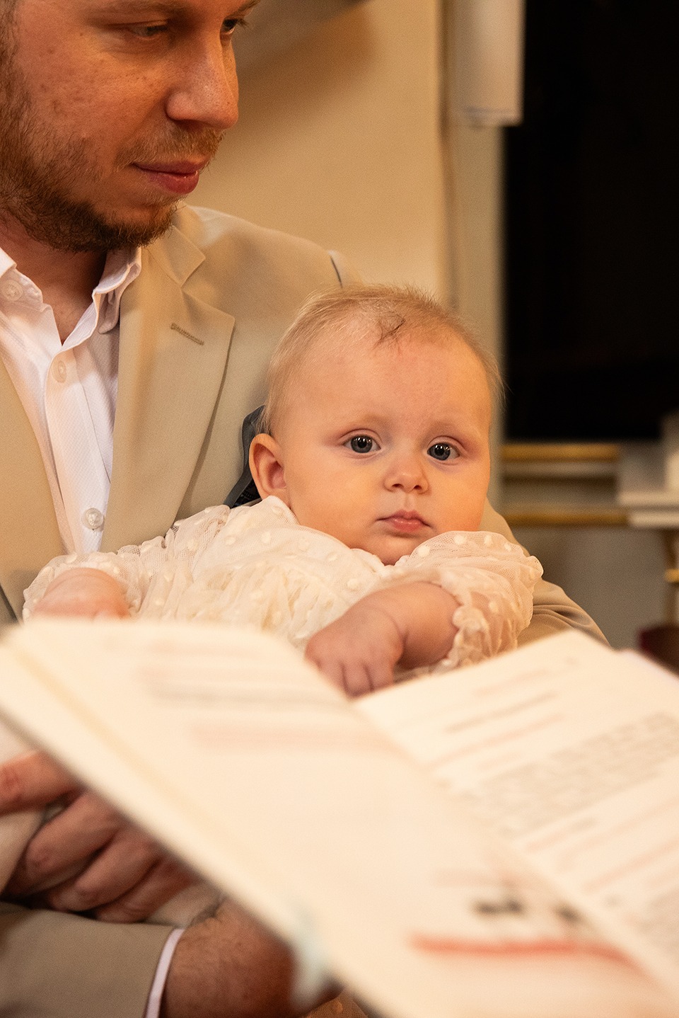 Urocze niemowlę w kremowej sukience, trzymane przez mężczyznę w beżowej marynarce, podczas lektury z otwartej księgi. Delikatne światło podkreśla detale.