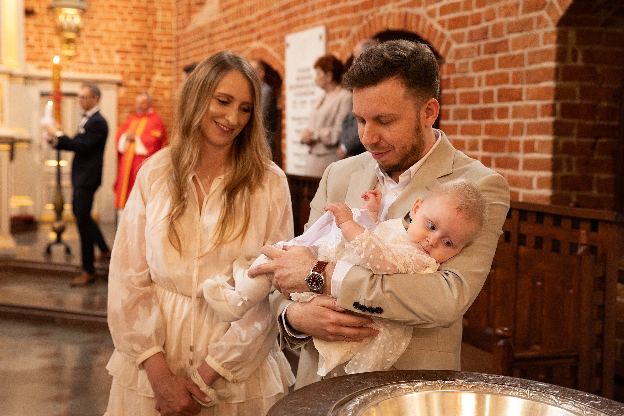 Rodzina w kościele podczas chrztu dziecka: ojciec w beżowym garniturze trzyma niemowlę w białej sukience, obok matka w jasnej sukience, ceglane ściany w tle.