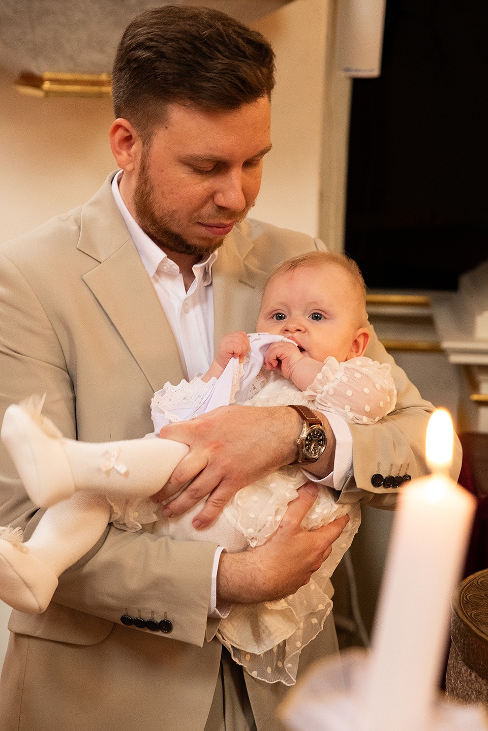 Portret ojca w beżowym garniturze trzymającego na rękach ubraną na biało dziewczynkę podczas ceremonii, w tle świeca.