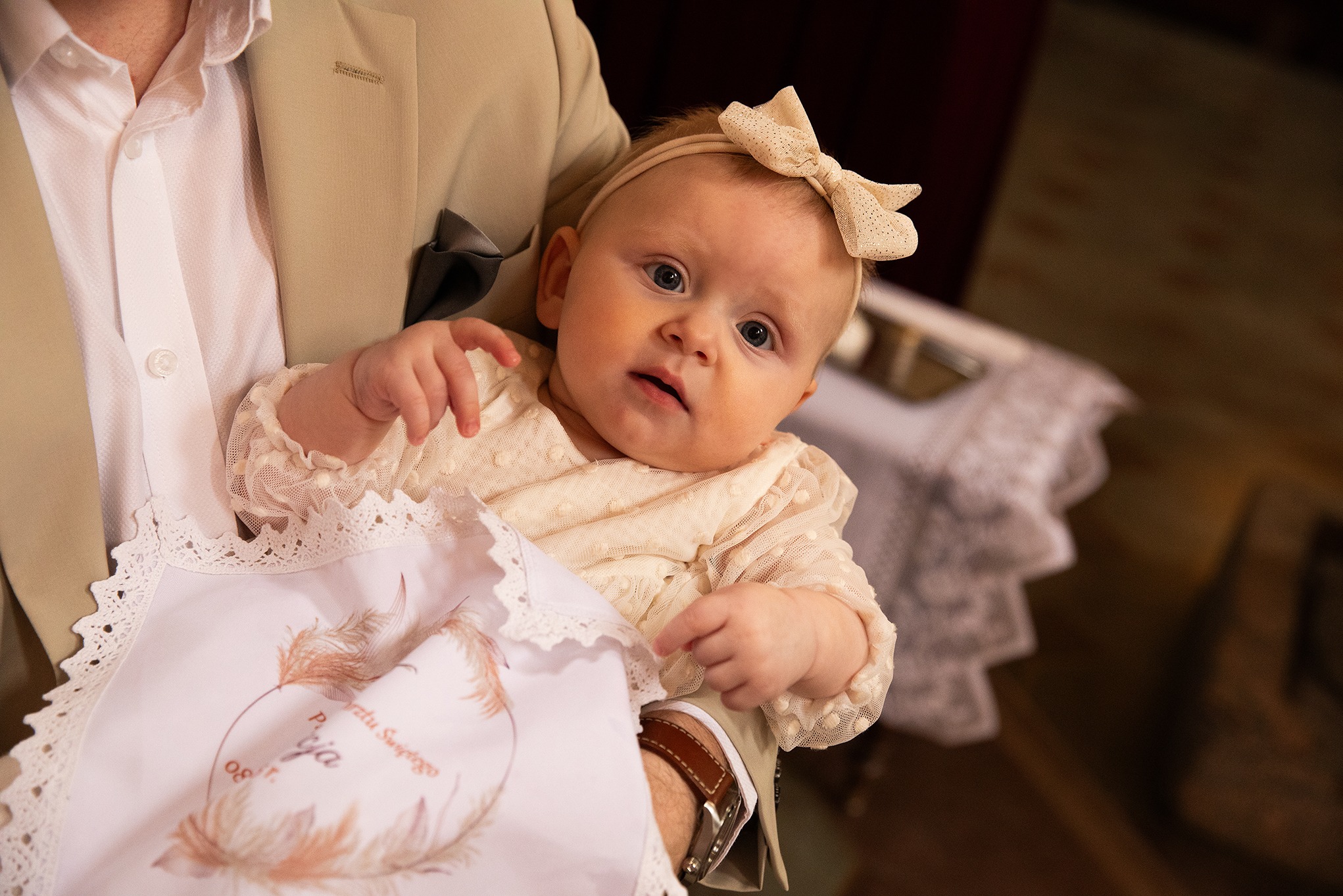 Urocze niemowlę w kremowej sukience i z kokardką na głowie, trzymane przez mężczyznę w beżowym garniturze, z widocznym haftowanym atłasowym kocykiem.