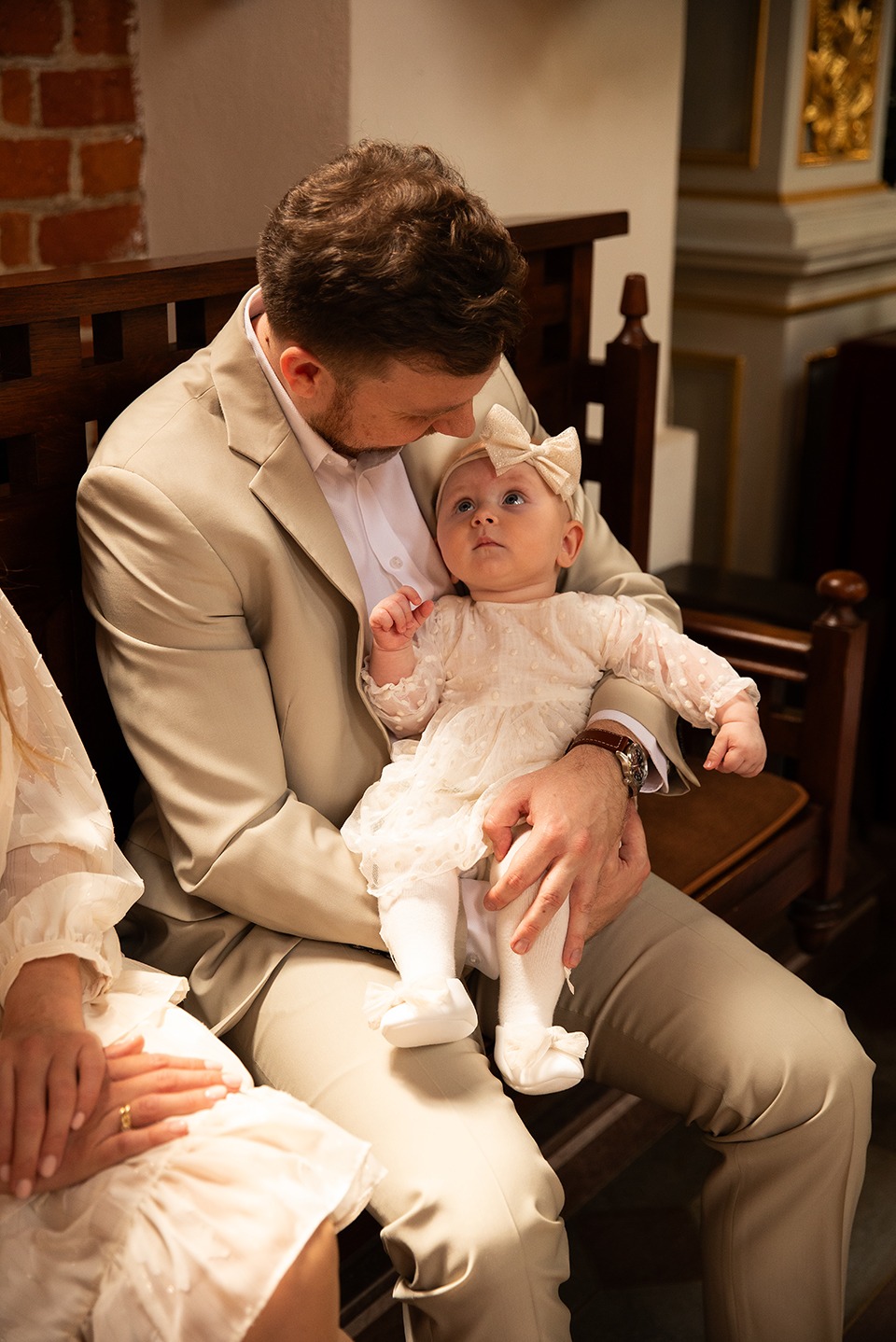 Ojciec w beżowym garniturze trzyma na kolanach uroczyste ubraną córeczkę w kościele. Dziewczynka patrzy w górę, jasne, stonowane kolory.