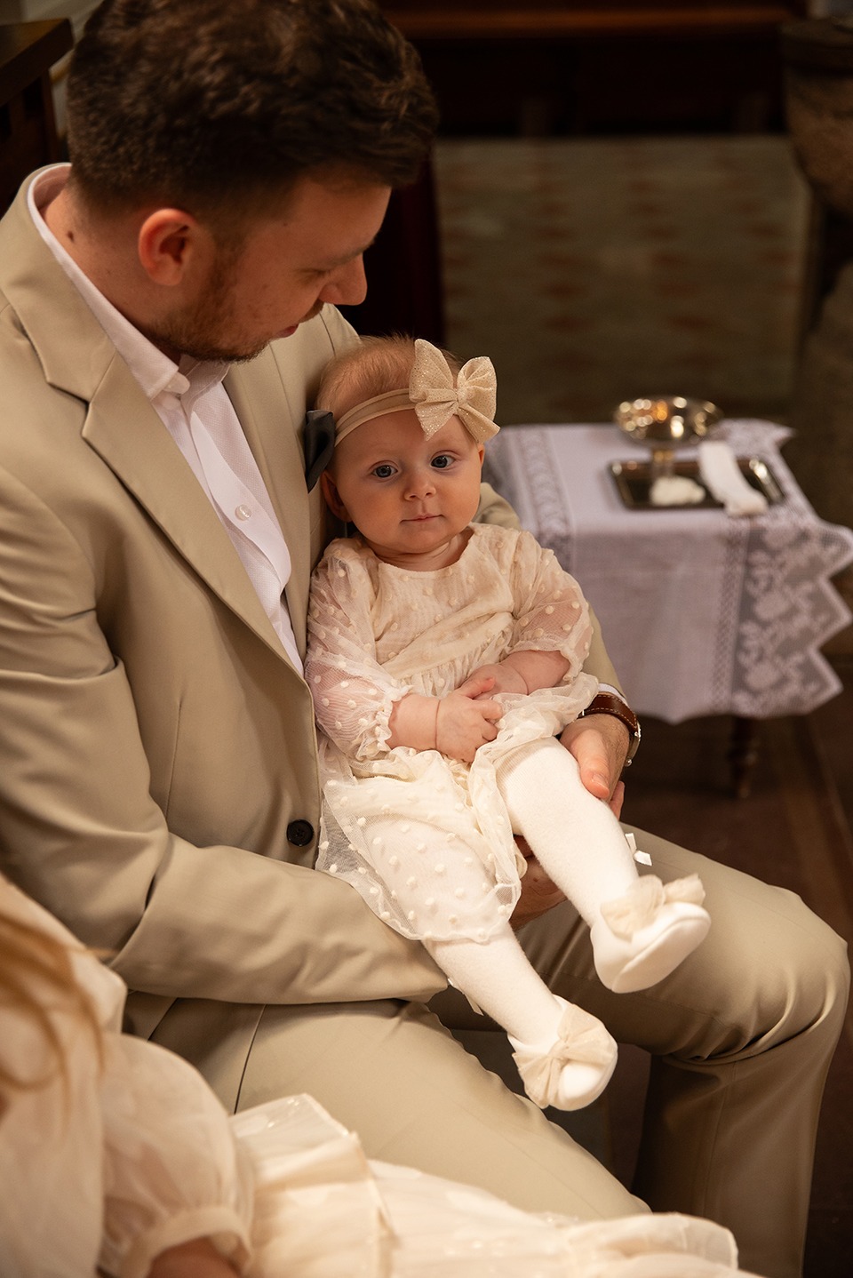 Ojciec w beżowym garniturze trzyma na kolanach niemowlę w kremowej sukience i opasce z kokardą, wnętrze kościoła, uroczystość chrztu.