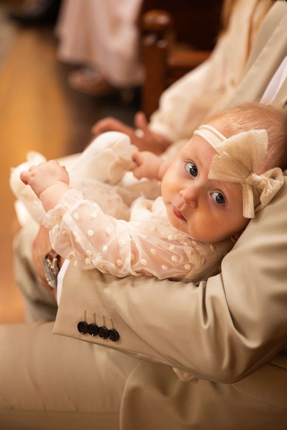 Niemowlę w kremowej sukience z kokardą na głowie, trzymane na rękach osoby w beżowym garniturze. Uwagę przykuwają duże, niebieskie oczy dziecka.