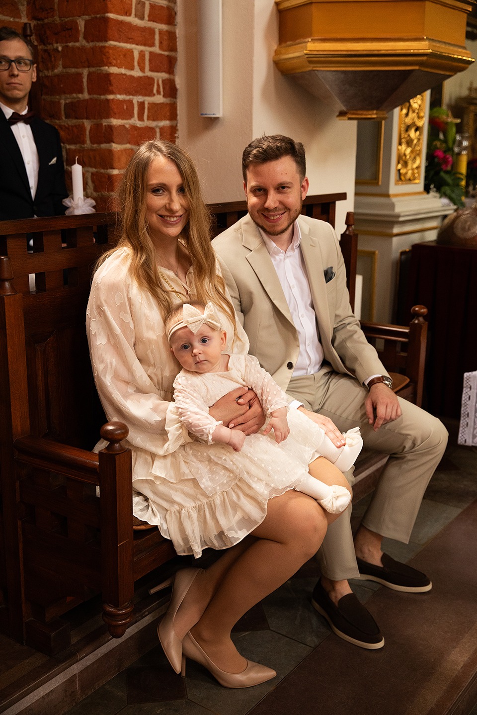 Uśmiechnięta rodzina siedzi w kościele, prawdopodobnie podczas uroczystości chrztu. Matka trzyma na kolanach ubraną na biało dziewczynkę z kokardą na głowie.