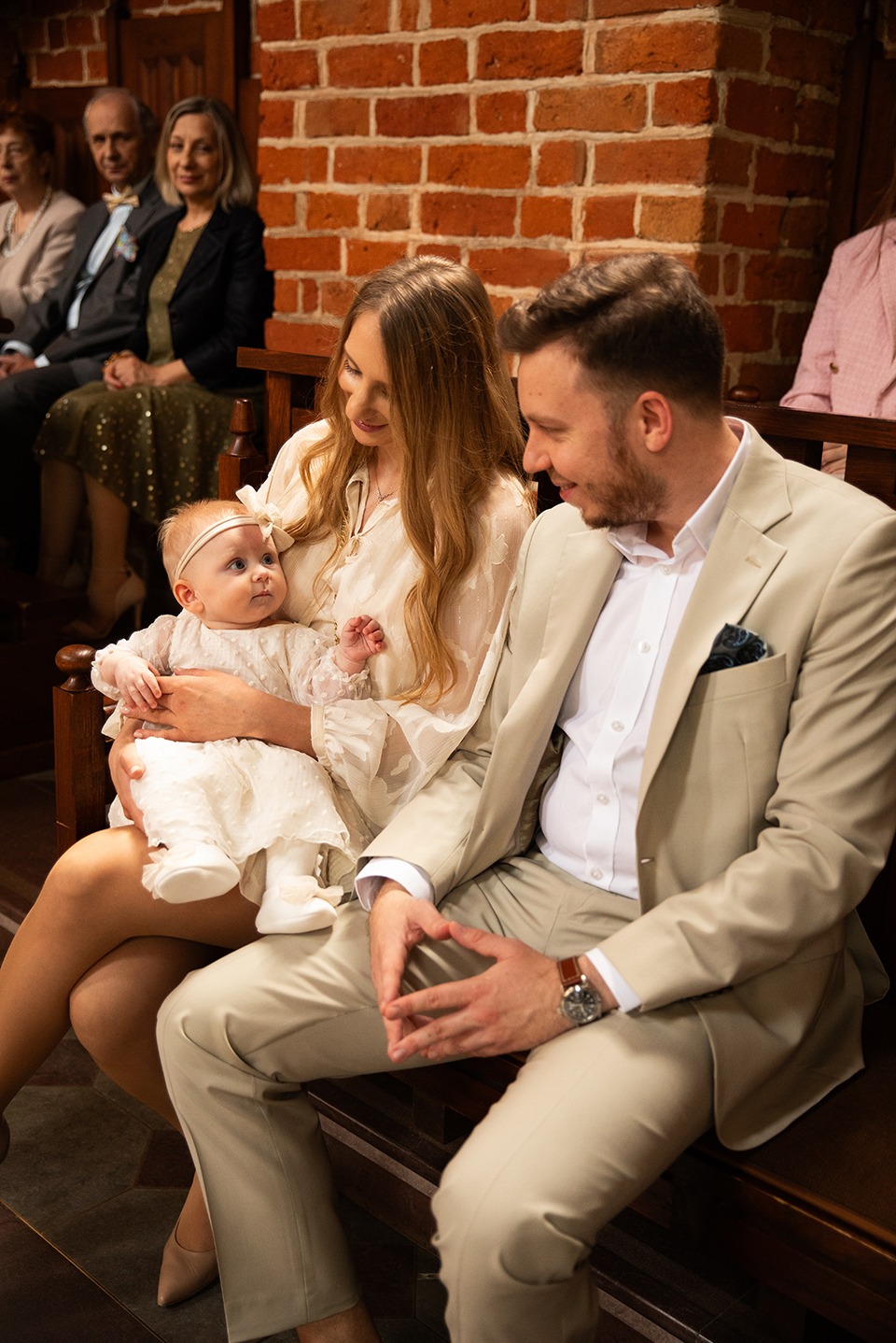 Uśmiechnięta rodzina w kościele: mama trzyma na kolanach niemowlę w białej sukience, obok tata w beżowym garniturze. W tle ceglana ściana i siedzący goście.