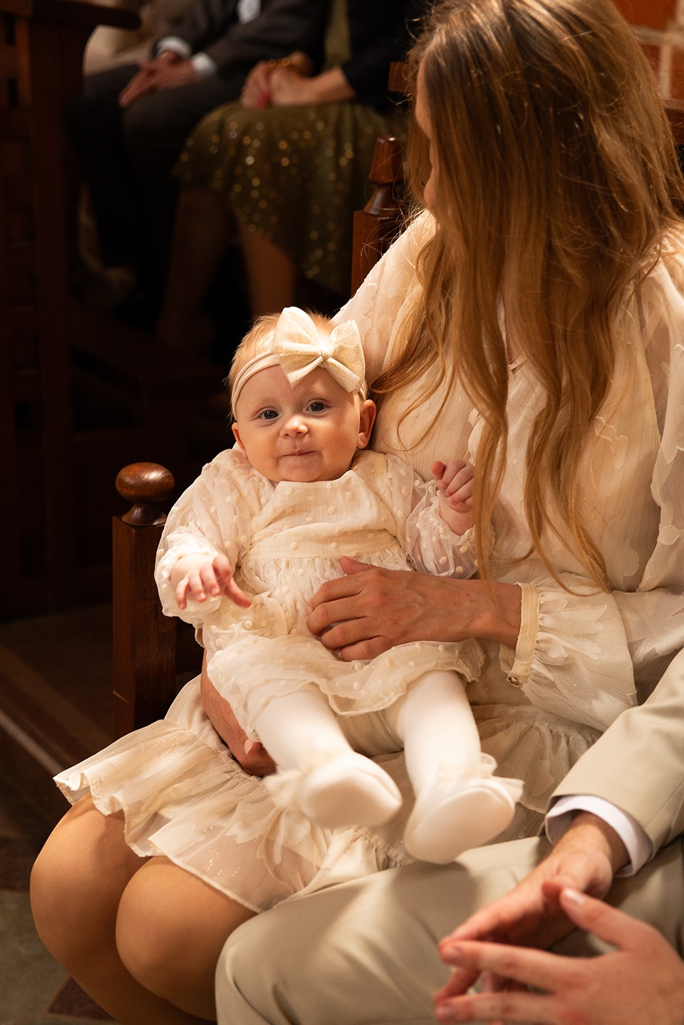 Urocza sesja: niemowlę w kremowej sukience i opasce z kokardką siedzi na kolanach matki w kościele, w tle widoczni goście.