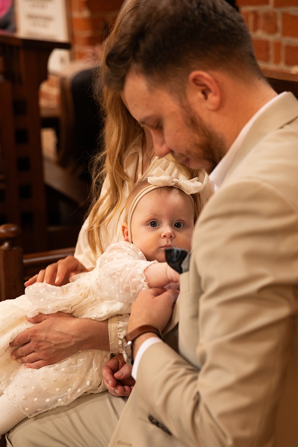 Ujęcie z chrztu: Ojciec w beżowym garniturze trzyma córeczkę w białej sukience z kokardką, Warszawa. Ciepłe światło, kościelne tło.