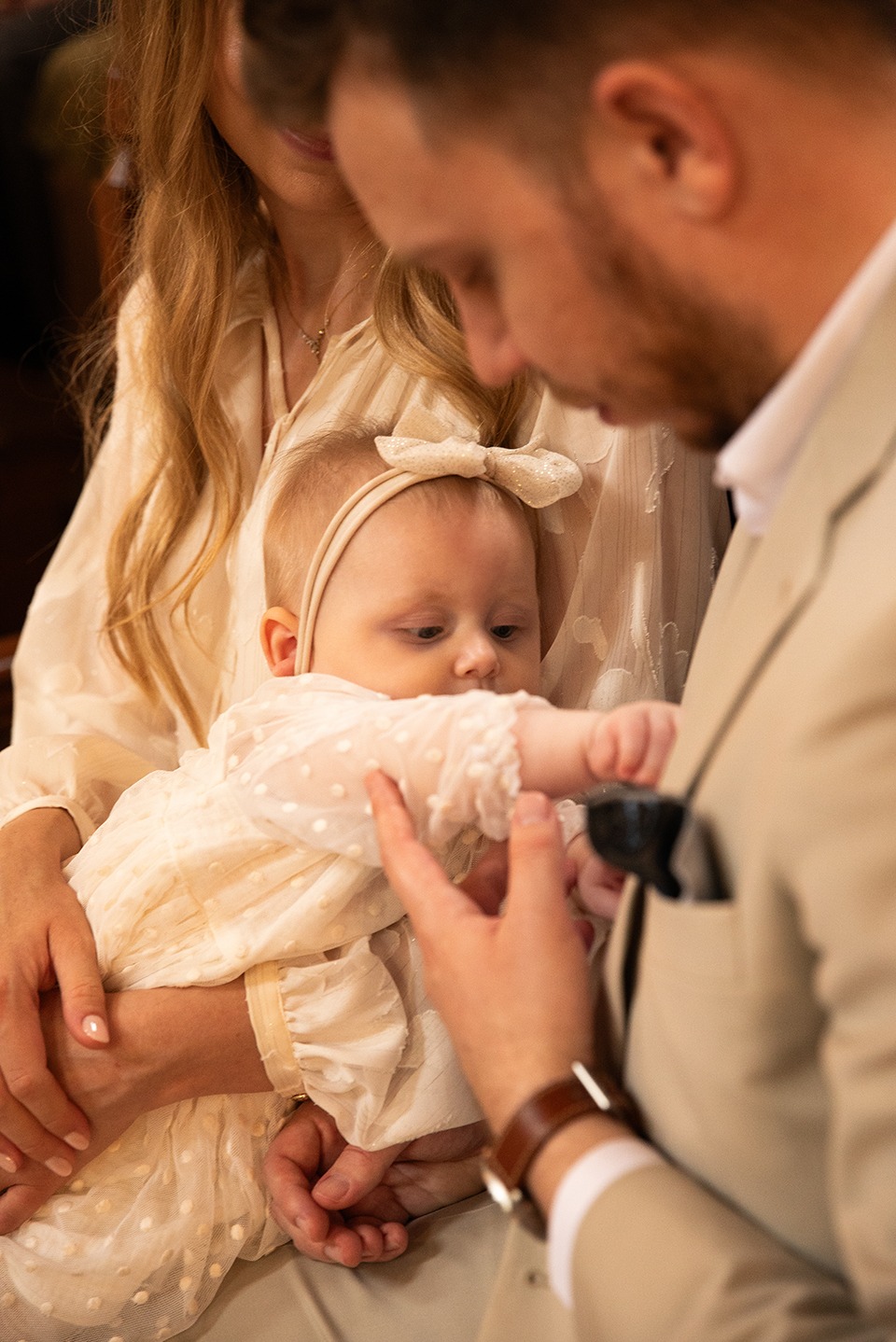 Dziecko w białej sukience z kokardą na głowie, trzymane przez rodziców, podczas ceremonii. Ujęcie z bliska, ciepłe oświetlenie, delikatne kolory.