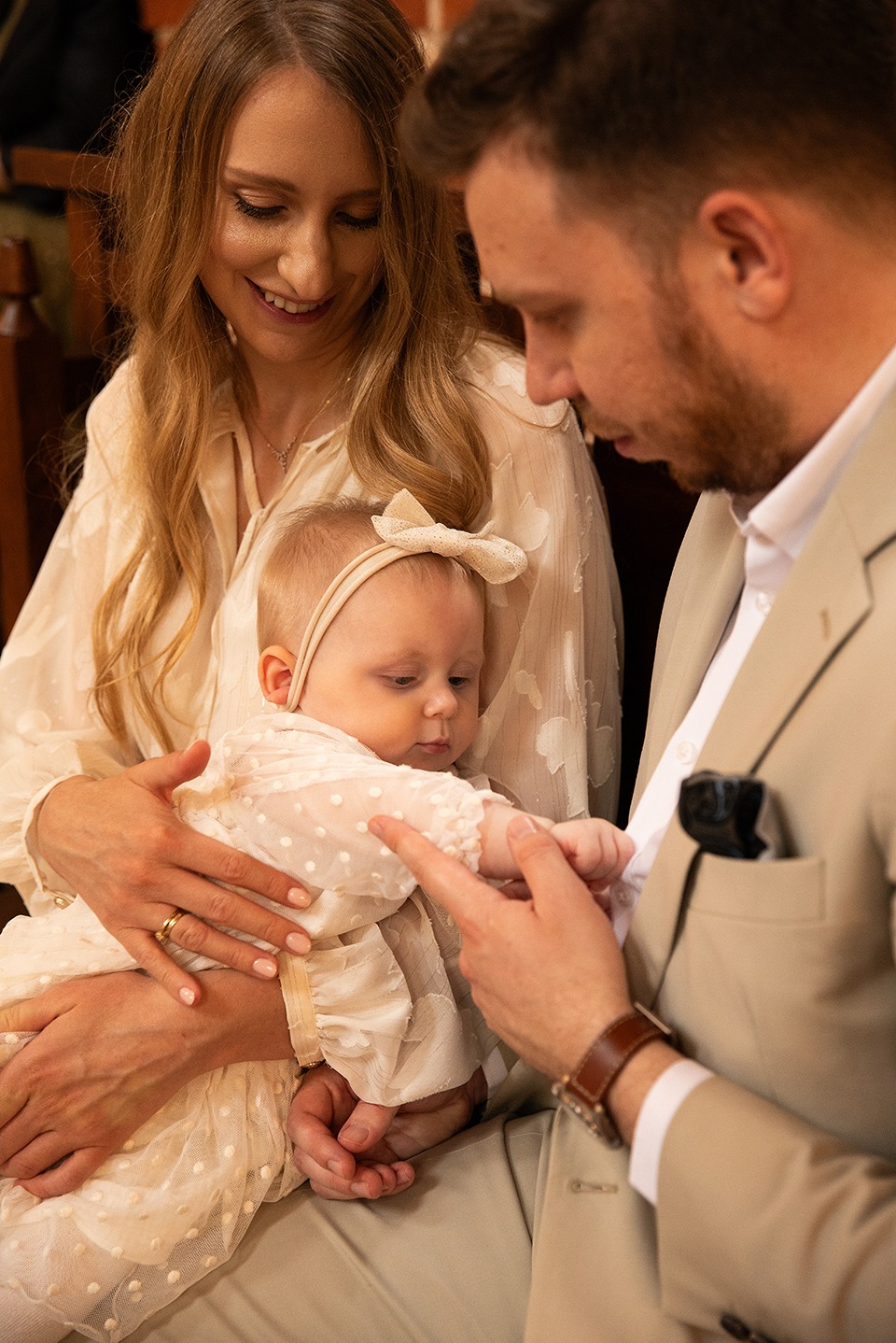 Uśmiechnięta mama trzyma na rękach niemowlę w kremowej sukience z kokardką, tata trzyma dziecko za rączkę. Jasne, naturalne światło, ciepłe kolory.