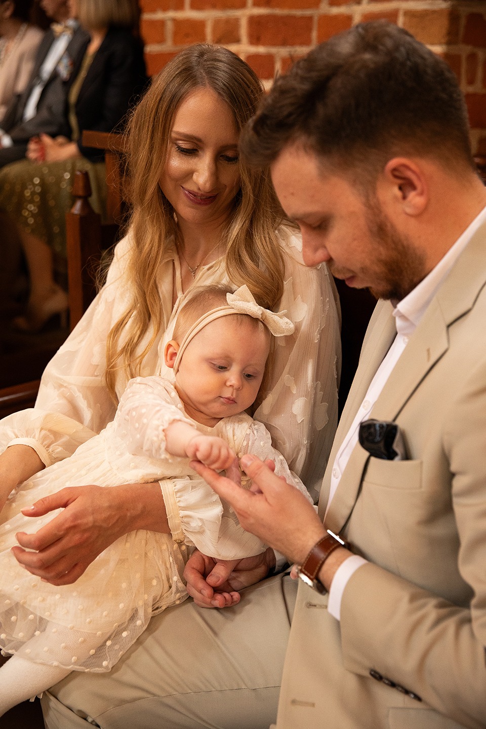 Rodzinne ujęcie z uroczystości chrztu: rodzice trzymają na kolanach niemowlę w kremowej sukience z kokardką, na tle ceglanej ściany i innych gości.