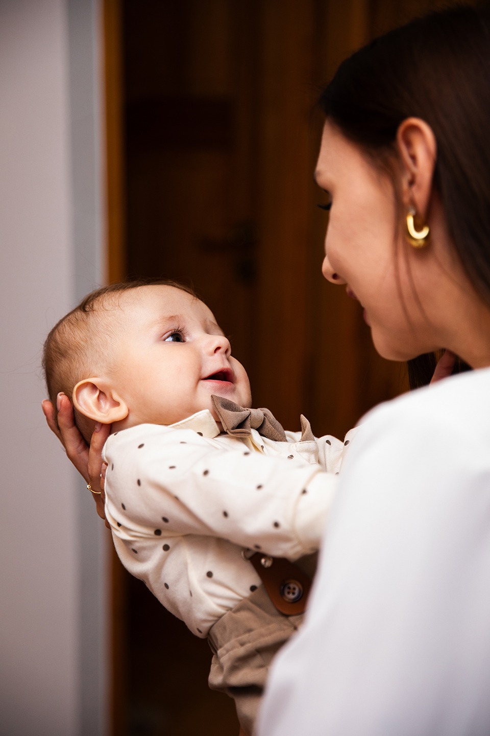 Uśmiechnięty niemowlak w eleganckiej koszuli z muchą, trzymany przez kobietę z kolczykami, patrzy w górę. Ciepłe, naturalne światło.