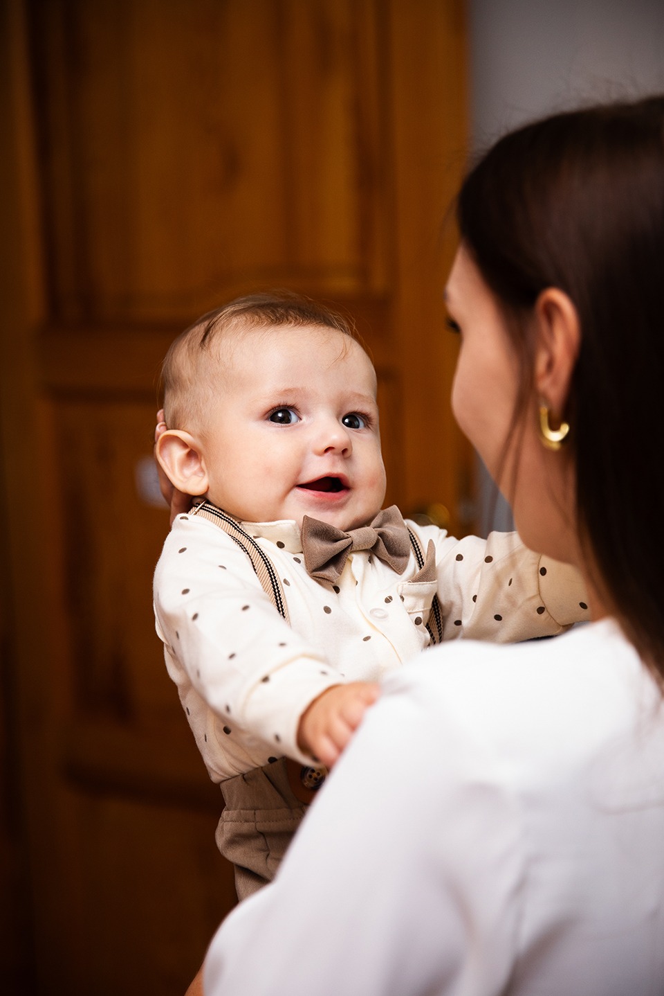 Uśmiechnięty niemowlak w eleganckim ubranku, z brązową muszką i szelkami, trzymany przez kobietę z kolczykami w kształcie kół. Ciepłe oświetlenie.