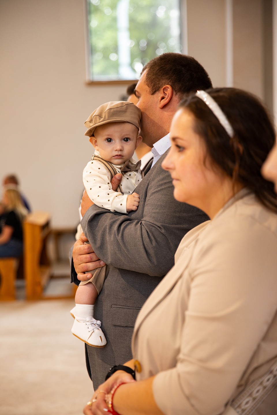 Uroczyste chrzciny: Ojciec trzyma elegancko ubranego synka w kaszkiecie, obok matka z opaską. W tle kościół i goście. Ciepłe, naturalne światło.