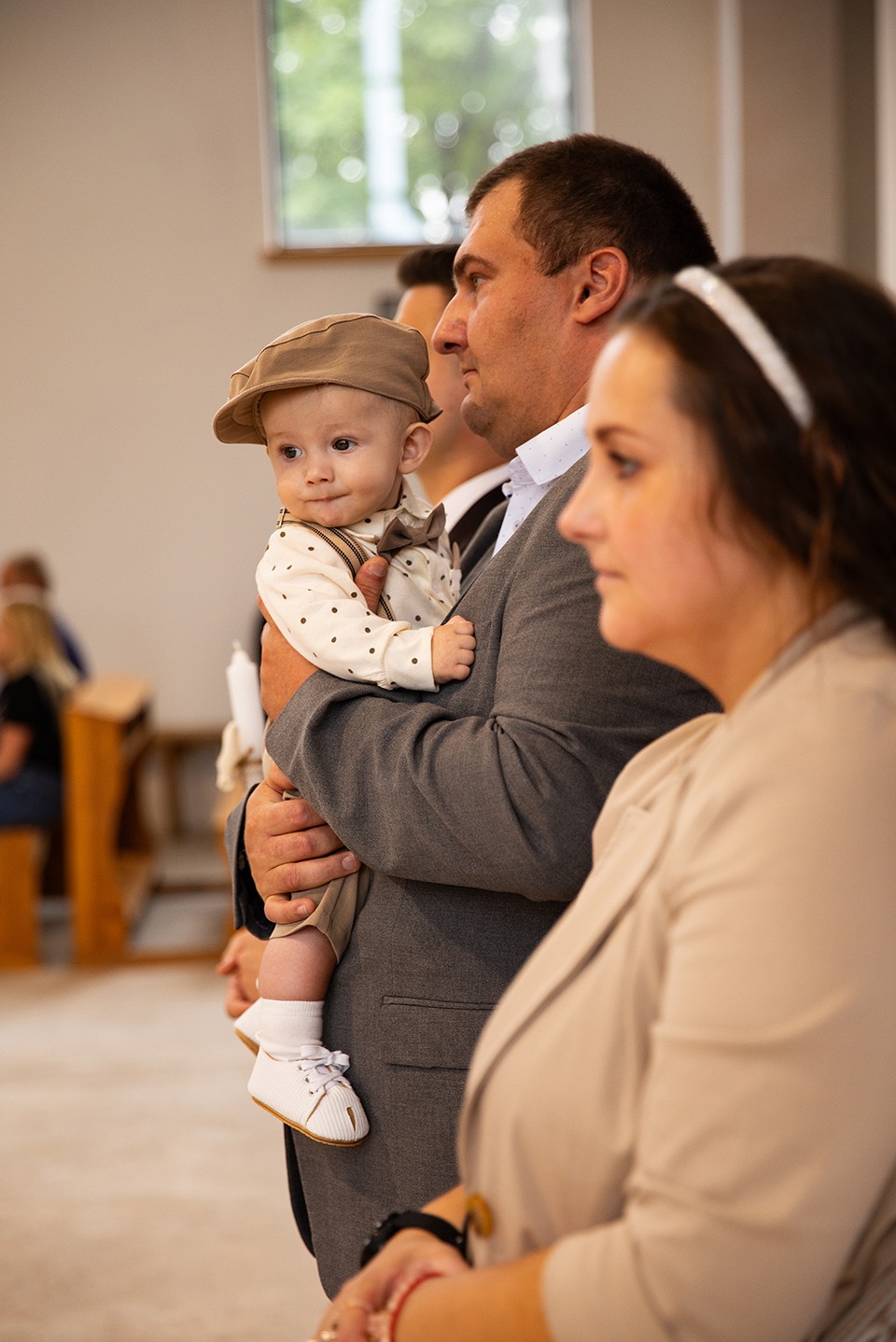 Uroczystość chrztu: Ojciec trzyma elegancko ubranego synka w kaszkiecie, obok matka. W tle widoczni goście w kościele. Delikatne, naturalne oświetlenie.