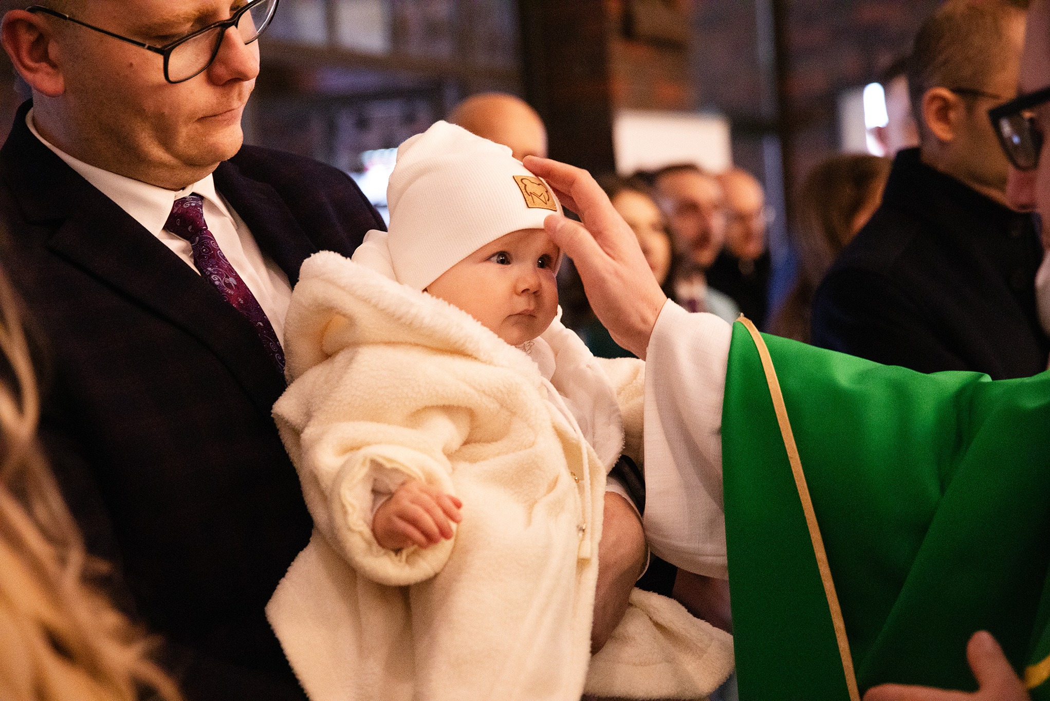 Chrzest dziecka w kościele: ksiądz dotyka głowy niemowlęcia w białym ubranku, trzymanego przez mężczyznę. W tle widoczni inni uczestnicy uroczystości.