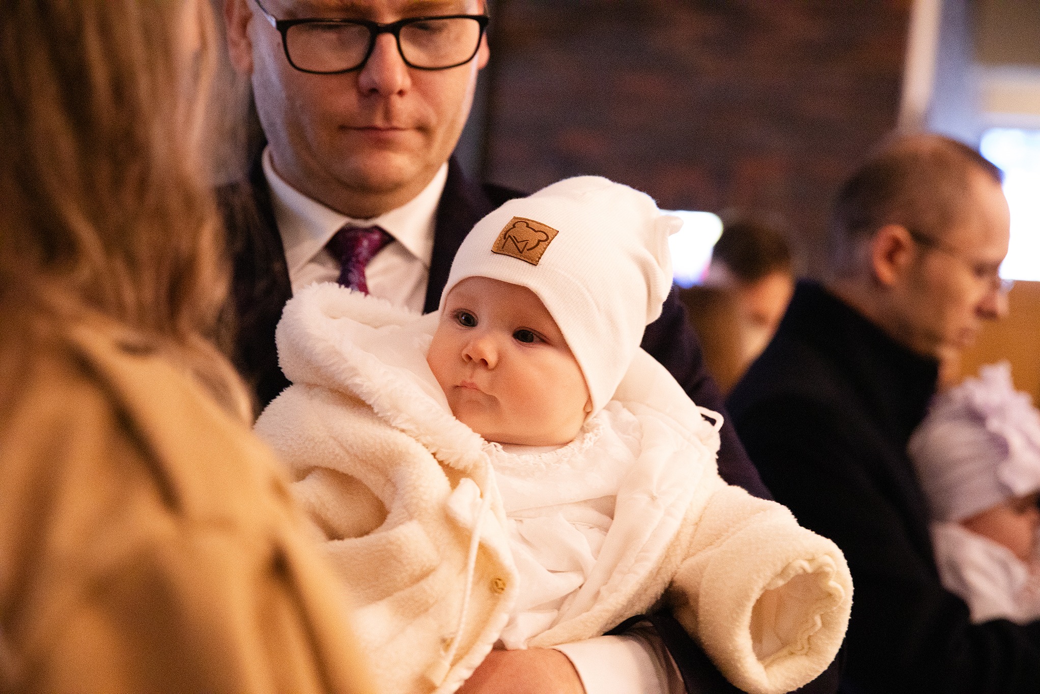 Urocze niemowlę w białym ubranku i czapeczce z misiem, trzymane przez mężczyznę w garniturze, w tle widoczni inni ludzie. Ciepłe, naturalne światło.