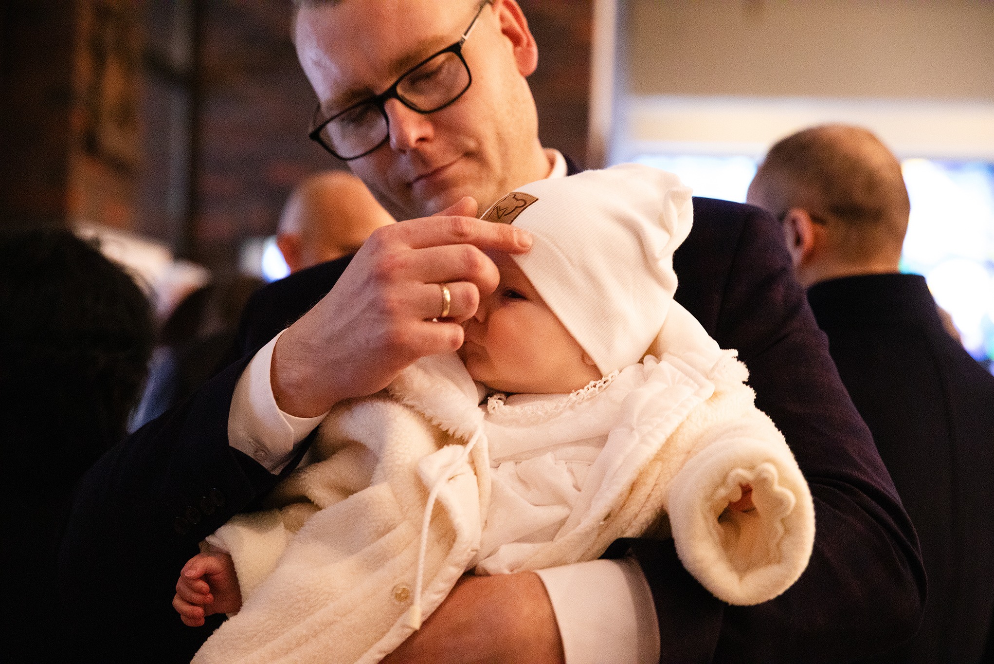 Ojciec w okularach czule poprawia czapeczkę śpiącemu niemowlęciu ubranemu w biały kombinezon. W tle widoczni inni ludzie, prawdopodobnie podczas uroczystości.