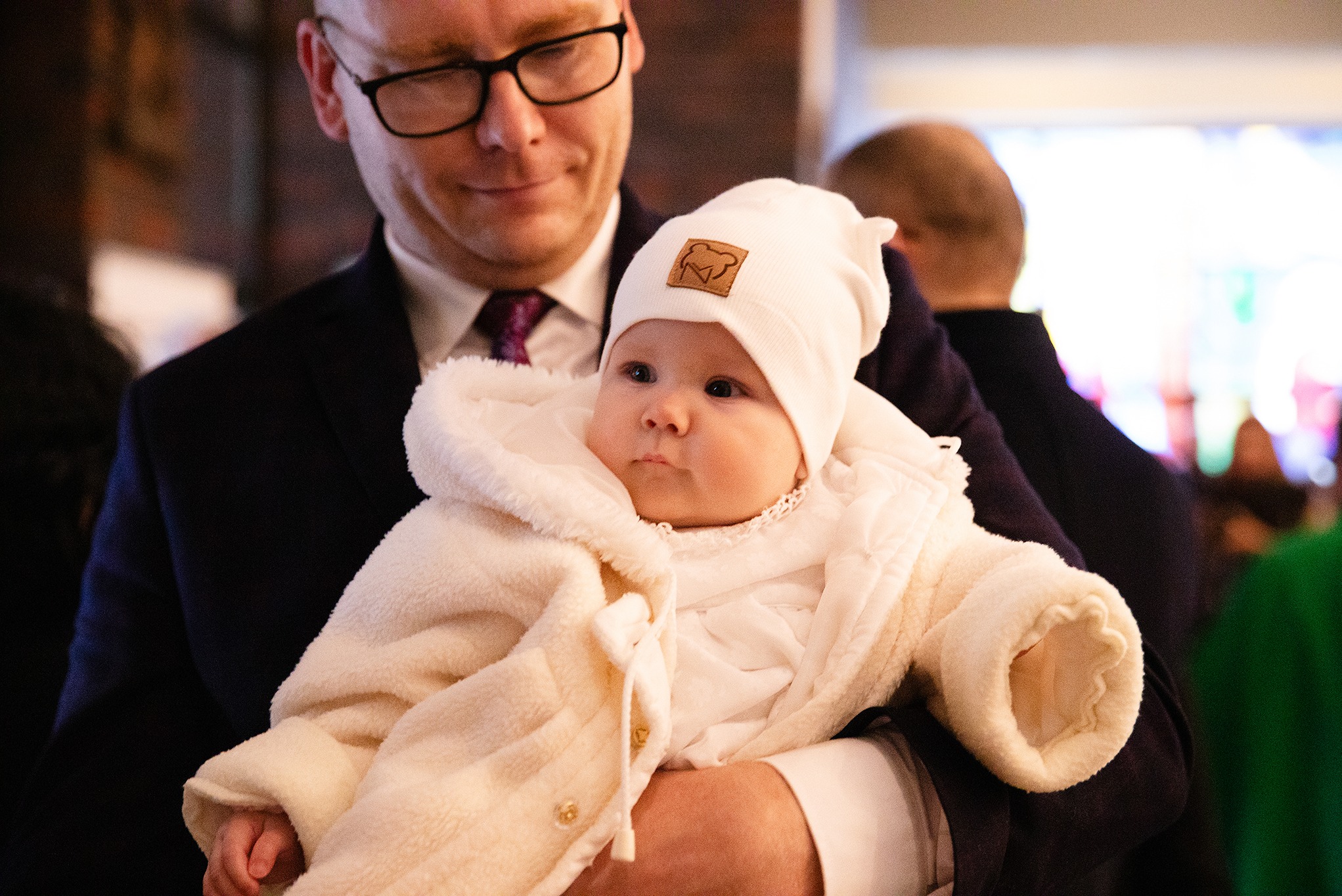 Urocze niemowlę w białym kombinezonie i czapce z naszywką, trzymane na rękach przez mężczyznę w garniturze, spogląda w bok z zaciekawieniem.