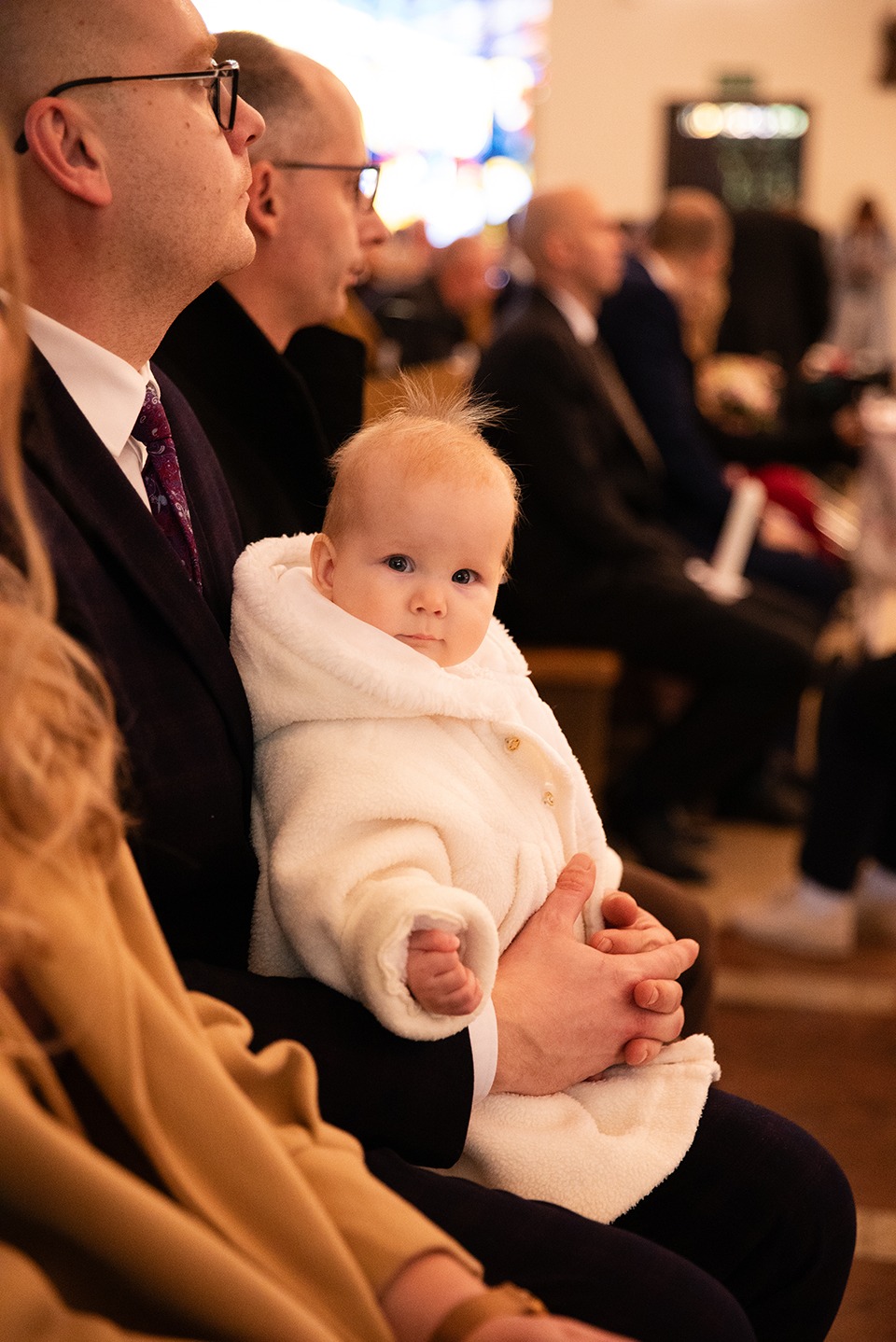 Urocze niemowlę w białym, pluszowym kombinezonie na kolanach mężczyzny podczas uroczystości w kościele, w tle widoczni inni goście.