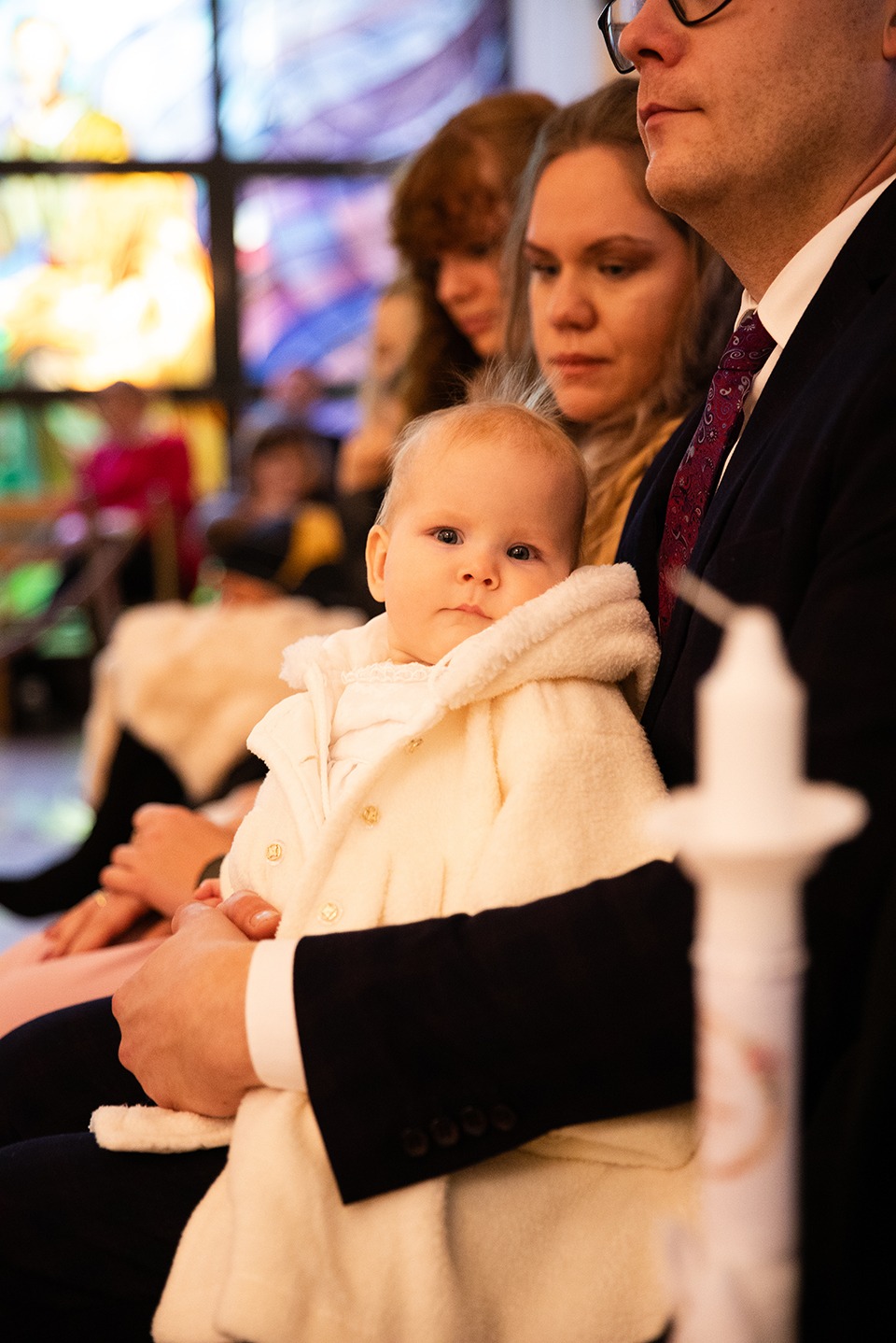 Ujęcie z uroczystości: niemowlę w kremowym ubranku na kolanach mężczyzny w garniturze, w tle witraże i skupione twarze rodziny. Świeca w pierwszym planie.