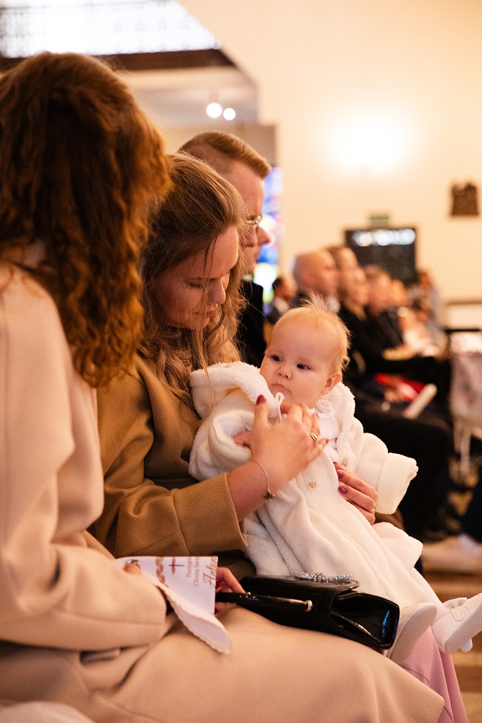 Matka trzyma na rękach niemowlę w białym ubranku podczas uroczystości w kościele. W tle widoczni inni uczestnicy wydarzenia.