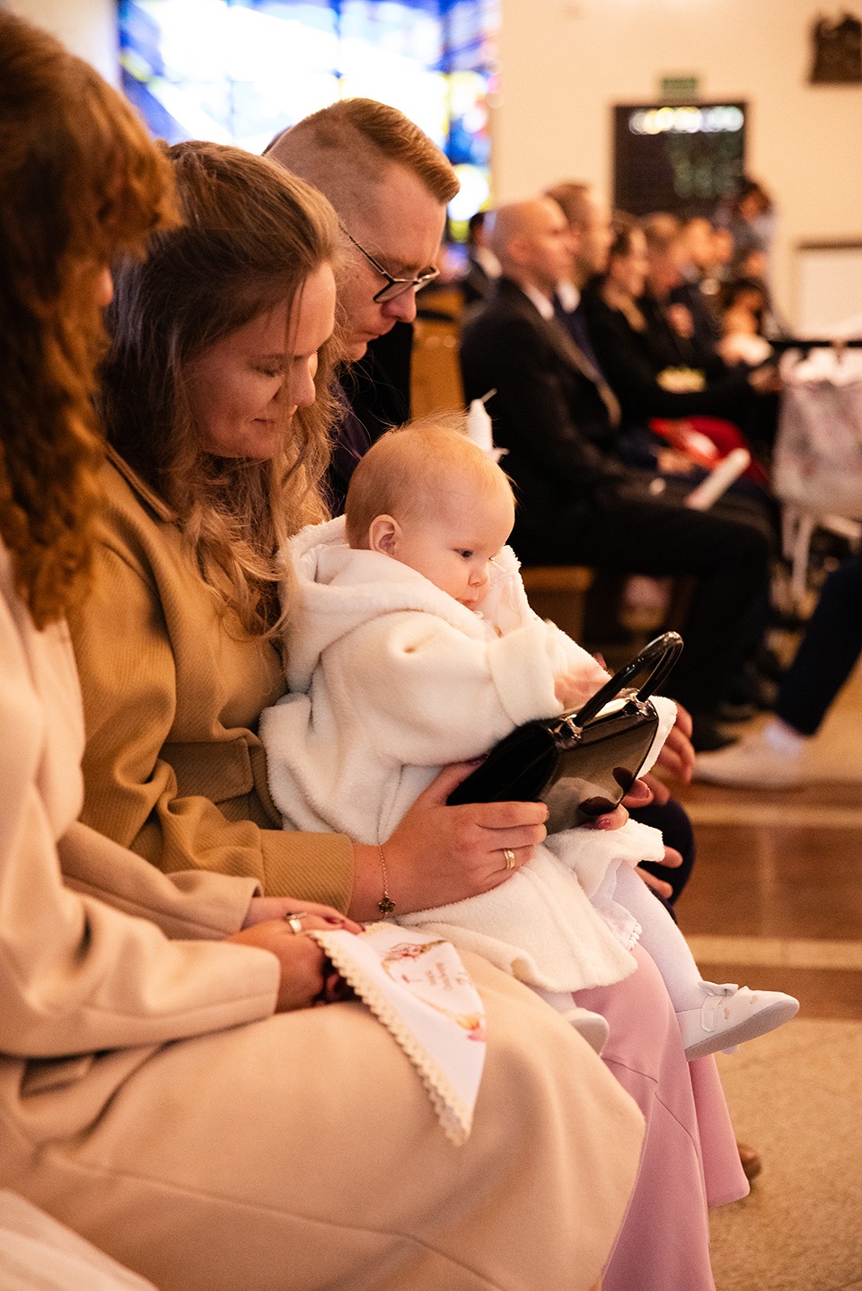 Rodzinne ujęcie w kościele: niemowlę w białym ubranku bawi się czarną torebką na kolanach mamy, w tle siedzący goście w stonowanych strojach.