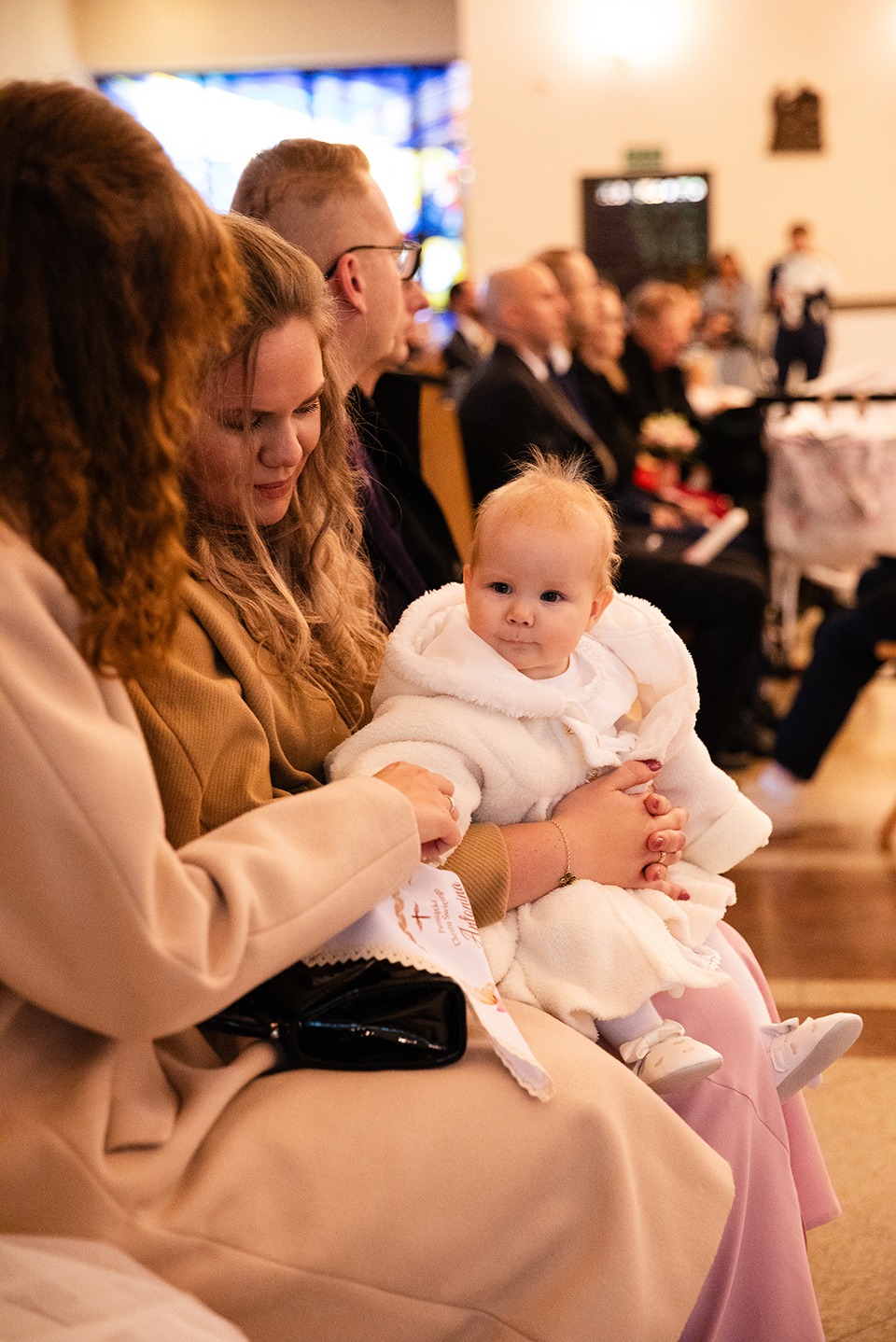 Uroczystość chrztu: niemowlę w białym ubranku na kolanach kobiety, w tle goście w kościele. W ręku kobiety pamiątka chrztu z imieniem dziecka.