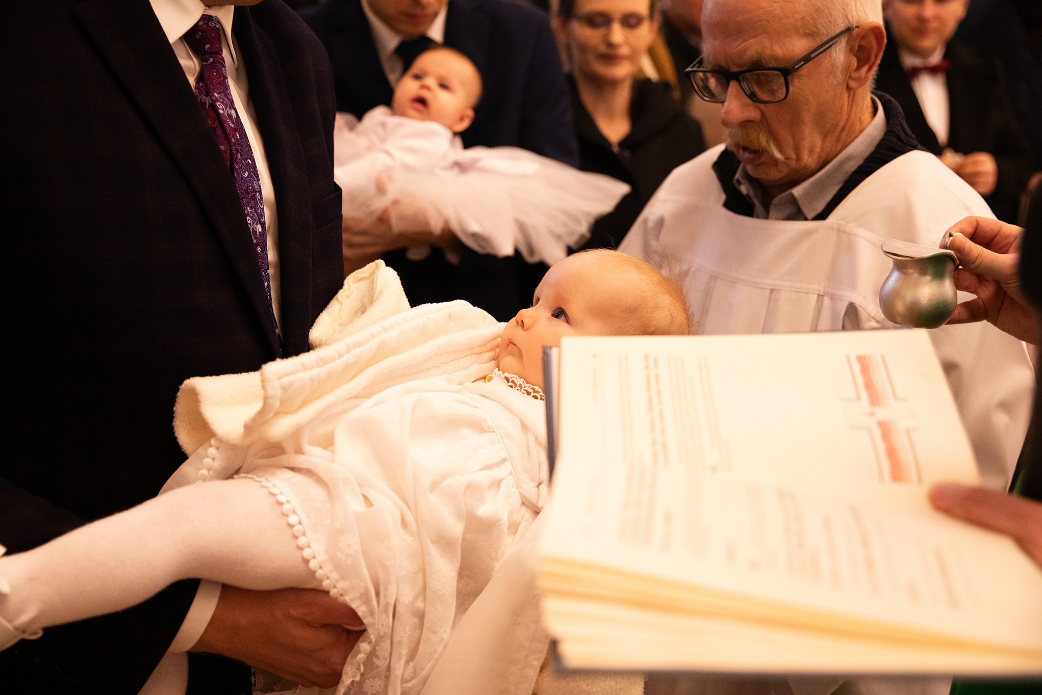 Chrzest dziecka: niemowlę w białej szacie trzymane przez mężczyznę, obok ksiądz z naczyniem i księgą, w tle inni goście i drugie dziecko. Uroczysta atmosfera.
