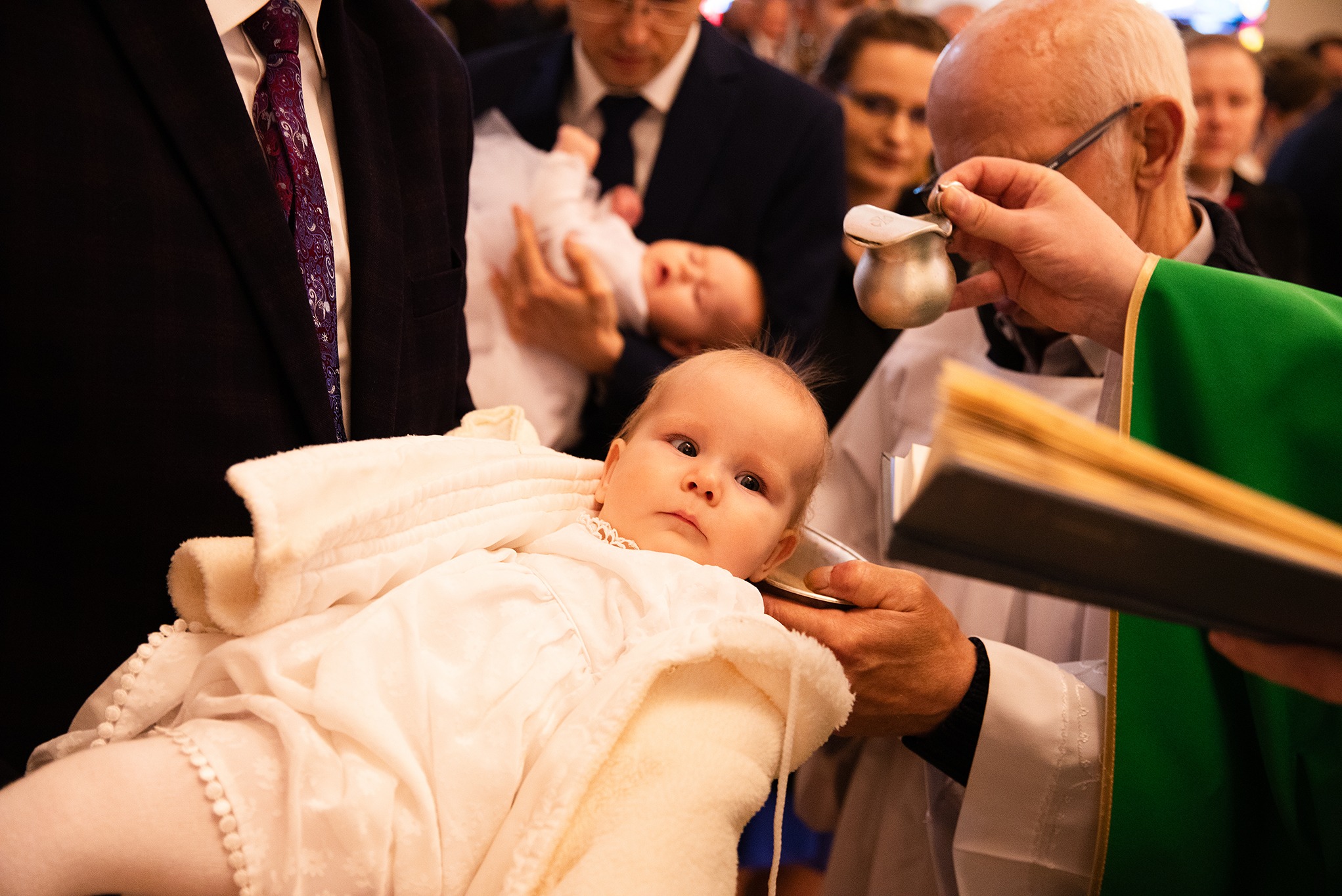 Ujęcie z ceremonii chrztu: niemowlę w białej szacie, trzymane przez osobę, nad nim ksiądz z naczyniem do chrztu i księgą, w tle rodzina.
