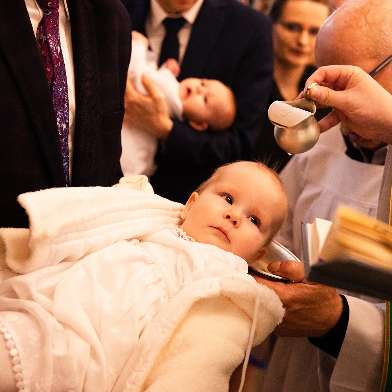 Ujęcie z ceremonii chrztu: niemowlę w białej szacie spoczywa na rękach, nad nim ksiądz z naczyniem do chrztu, w tle rodzina. Ciepłe, stonowane światło.