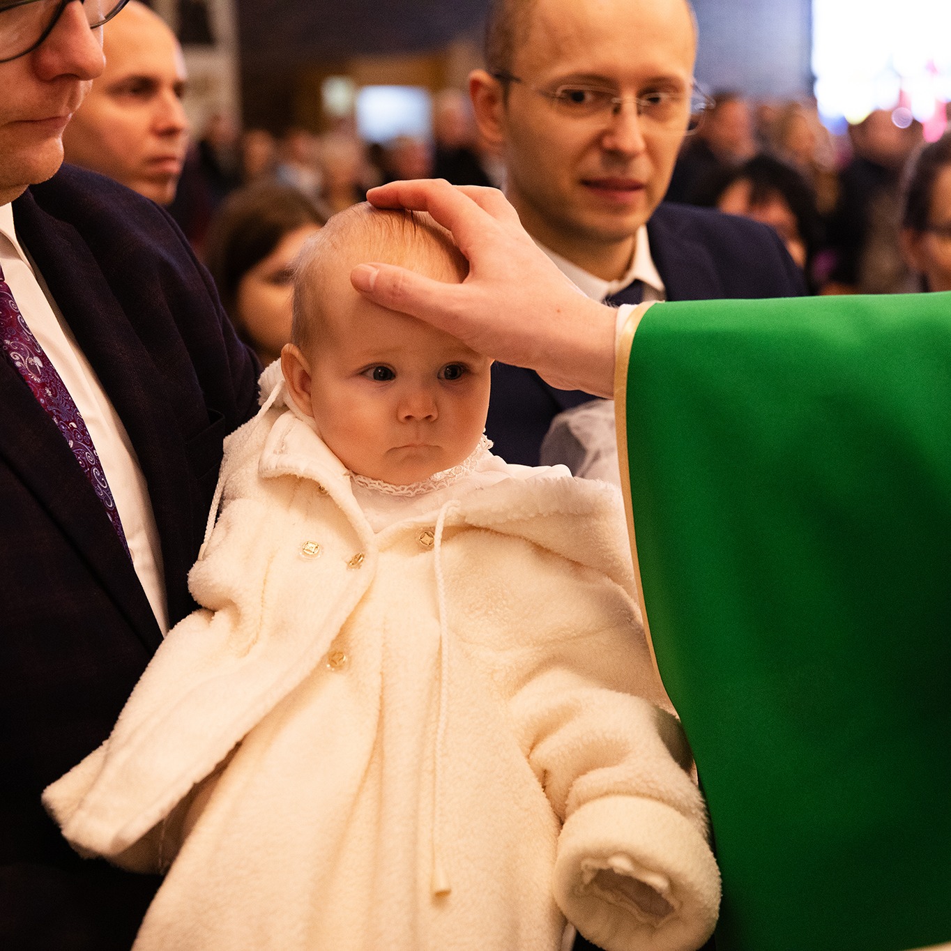 Ksiądz błogosławi niemowlę w białym ubranku podczas ceremonii w kościele, w tle widoczni goście. Uroczystość religijna, skupienie na dziecku i duchownym.