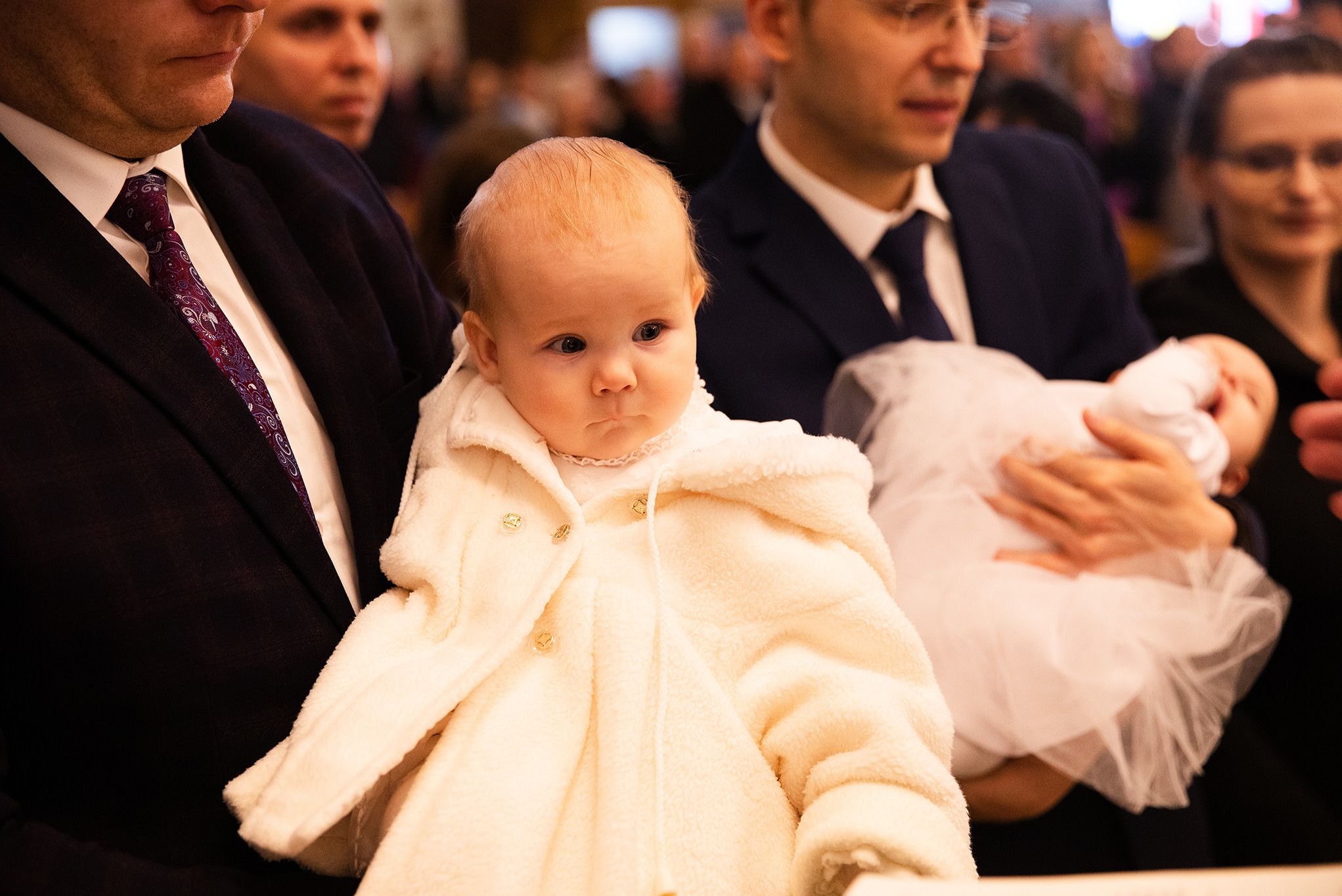 Urocze niemowlę w kremowym, pluszowym okryciu na rękach mężczyzny w garniturze podczas uroczystości. W tle widoczni inni goście i kolejne dziecko.
