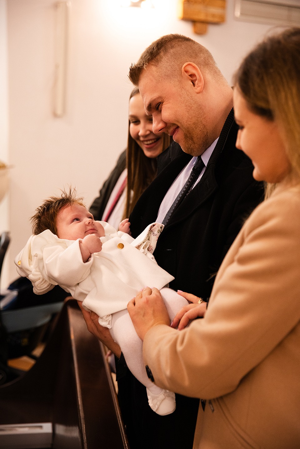Uśmiechnięty ojciec trzyma na rękach niemowlę w białym ubranku, w tle dwie kobiety obserwują scenę w kościele. Ciepłe oświetlenie podkreśla rodzinny charakter.
