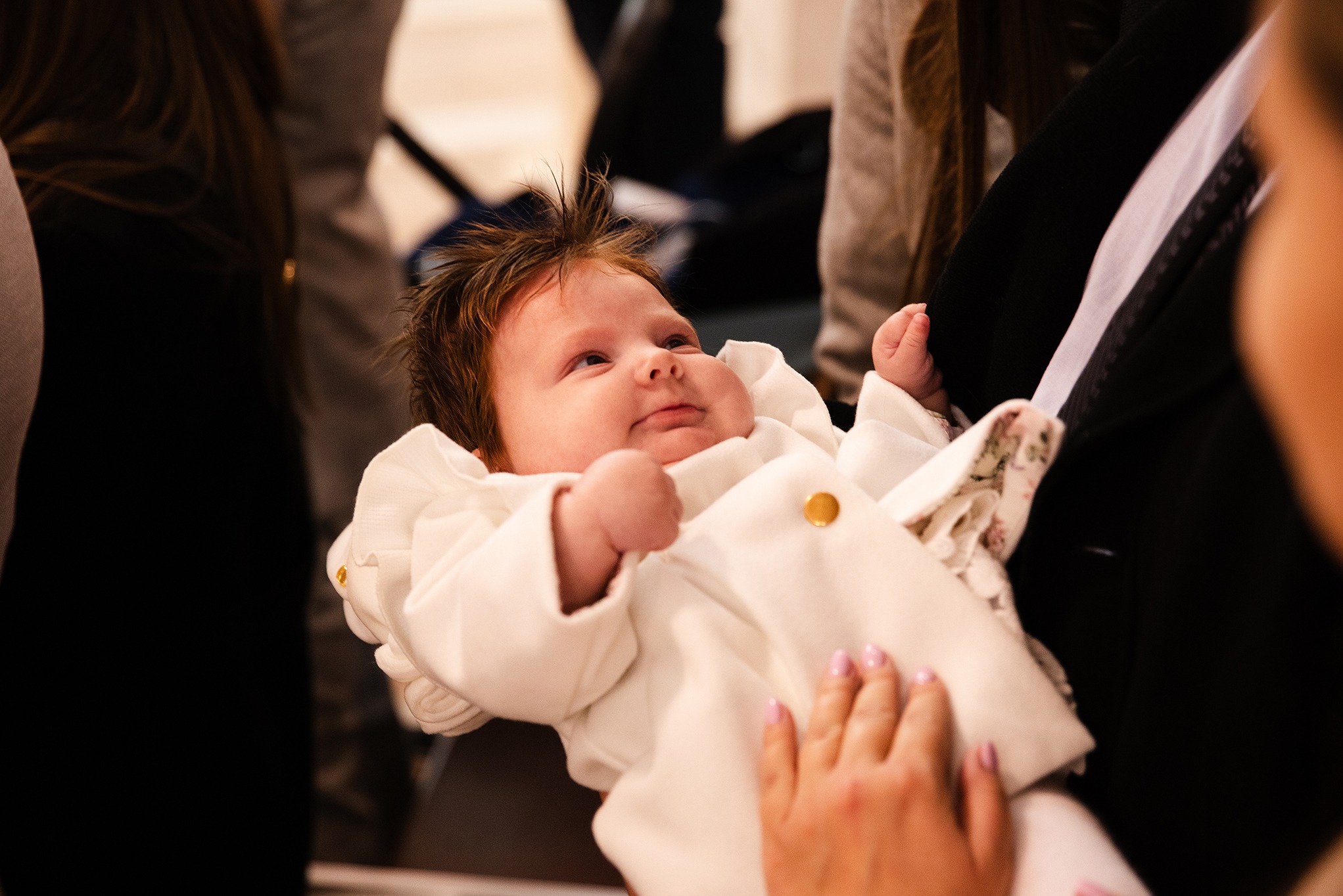 Urocze niemowlę w białym ubranku, z delikatnym uśmiechem, trzymane na rękach. Widoczne dłonie osoby trzymającej, miękkie światło podkreśla detale.